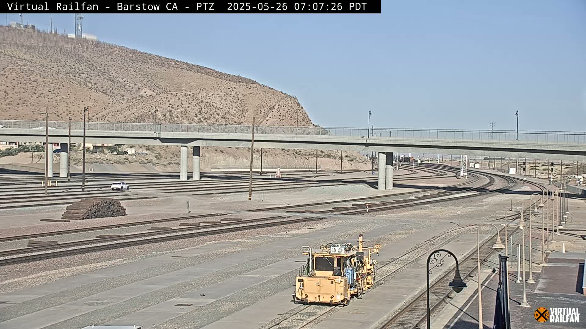 A yellow railway maintenance vehicle sits on a set of train tracks under a bridge on a sunny day, with a desert landscape and numerous other tracks extending into the distance.