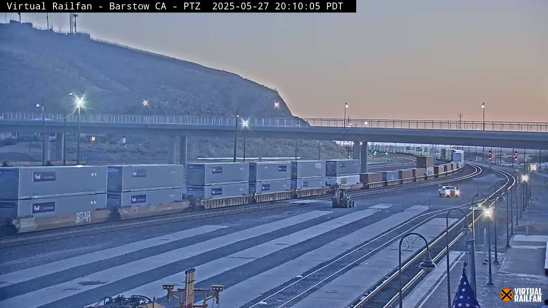 A long freight train carrying multiple shipping containers passes under a bridge at dusk.