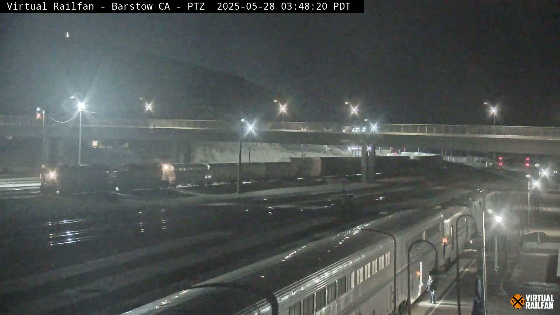 A long passenger train is parked at night at a station under a highway overpass, with some snow visible on the ground.