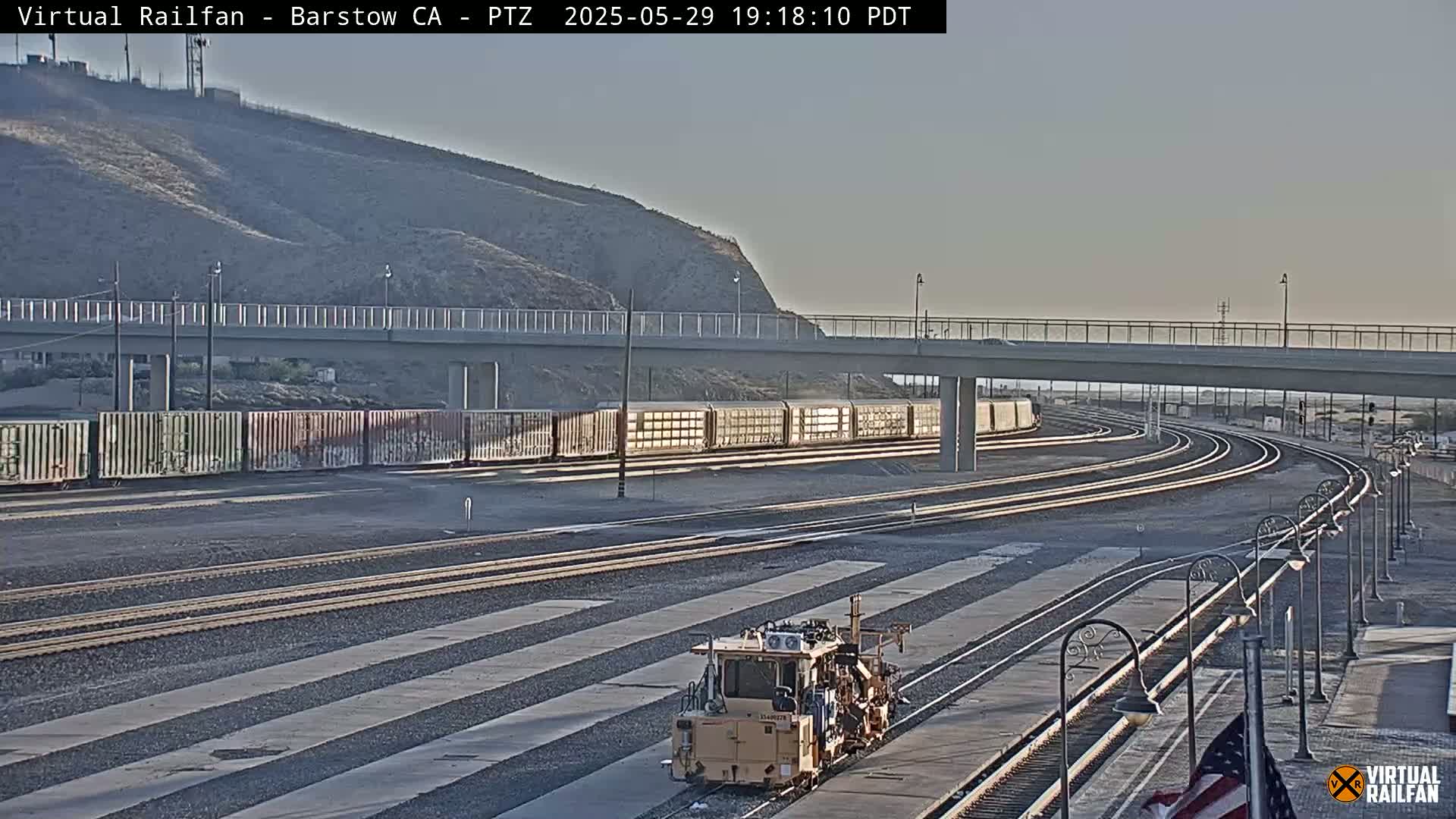 Under a clear sky, a rail yard with a train and railway maintenance vehicle is visible, with a road overpass in the background.