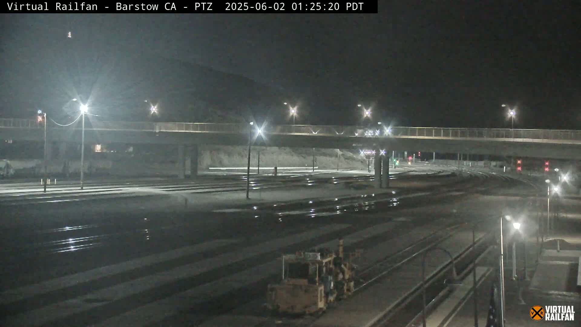 A nighttime view of a railway yard with a road overpass, illuminated by streetlights, under a clear, dark sky.