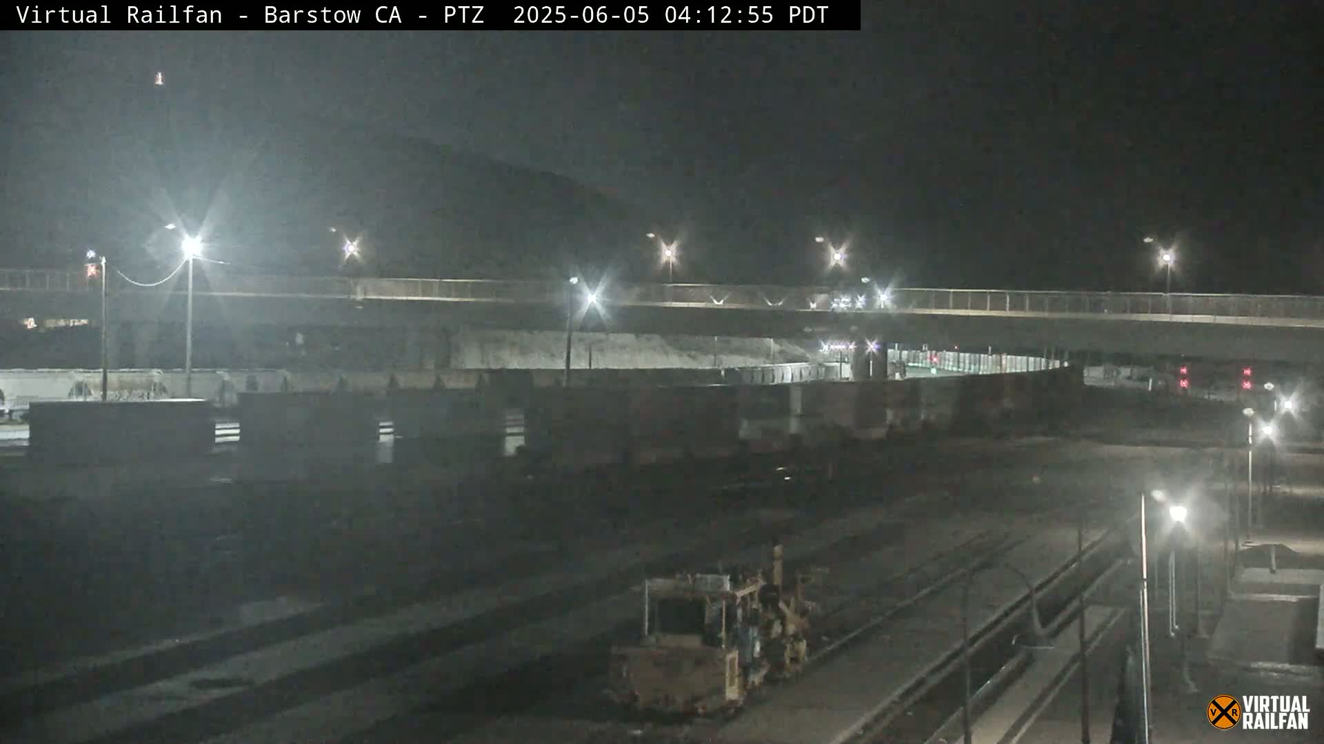 A nighttime view of a rail yard with a highway overpass, illuminated by streetlights, under a clear sky.