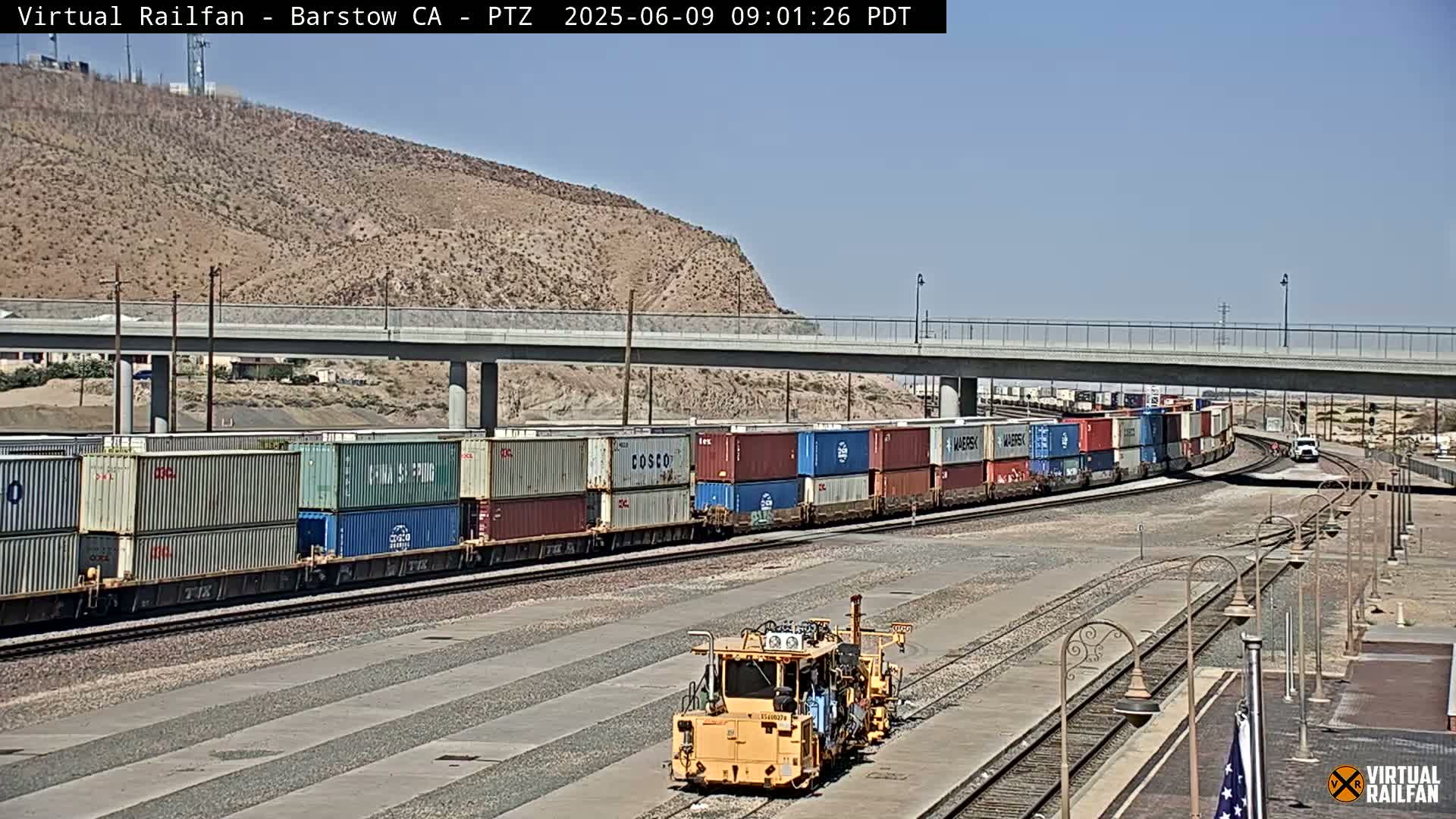 A long freight train carrying numerous shipping containers passes under a highway overpass on a sunny day.