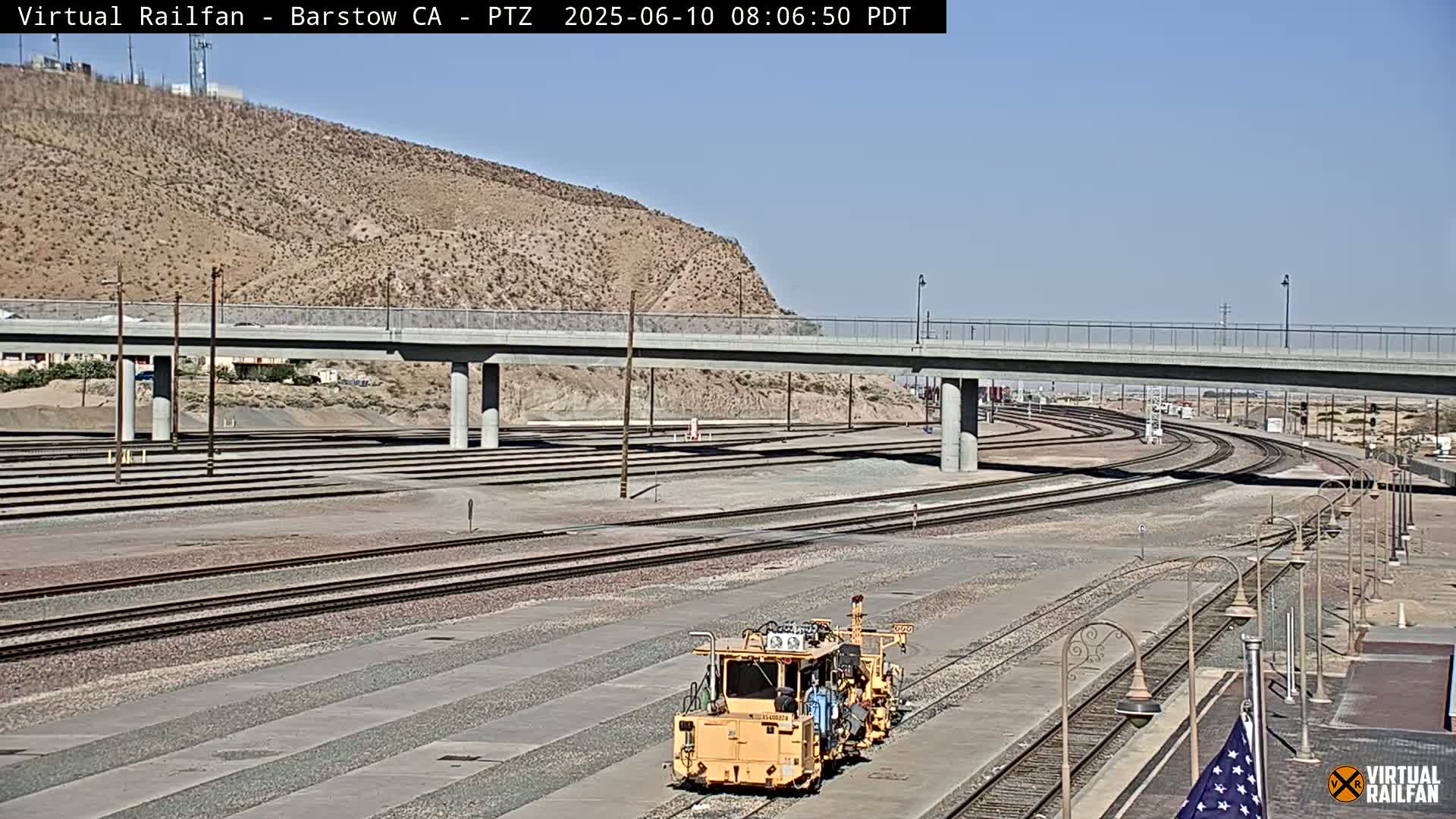 A yellow railway maintenance vehicle sits on a network of train tracks that curve under a bridge on a clear, sunny day.