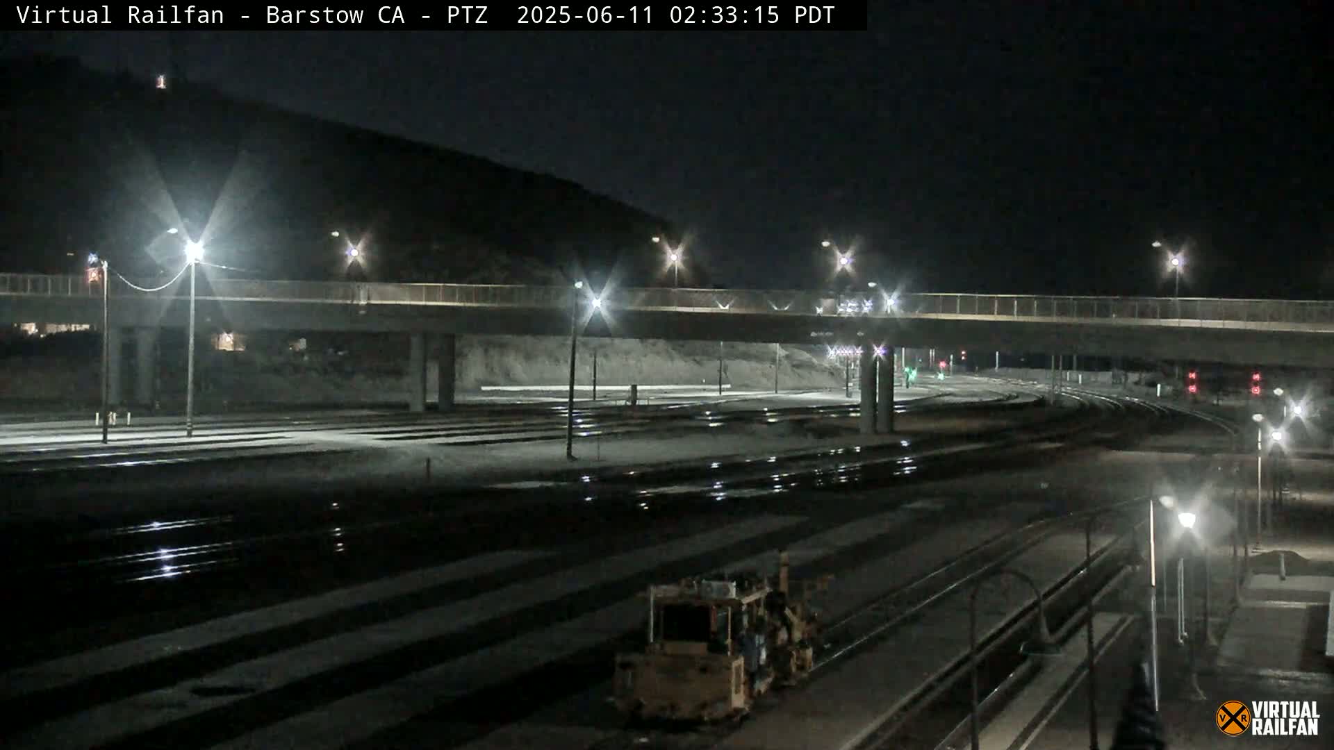 A nighttime view of a railway yard with a roadway overpass, lit by streetlights, shows a piece of railway maintenance equipment near the tracks.