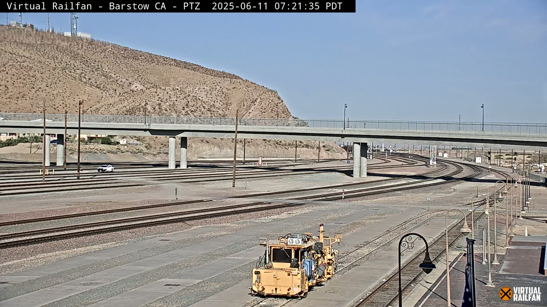 A rail yard with multiple train tracks under a bridge on a sunny day, and a small yellow maintenance vehicle is visible in the foreground.