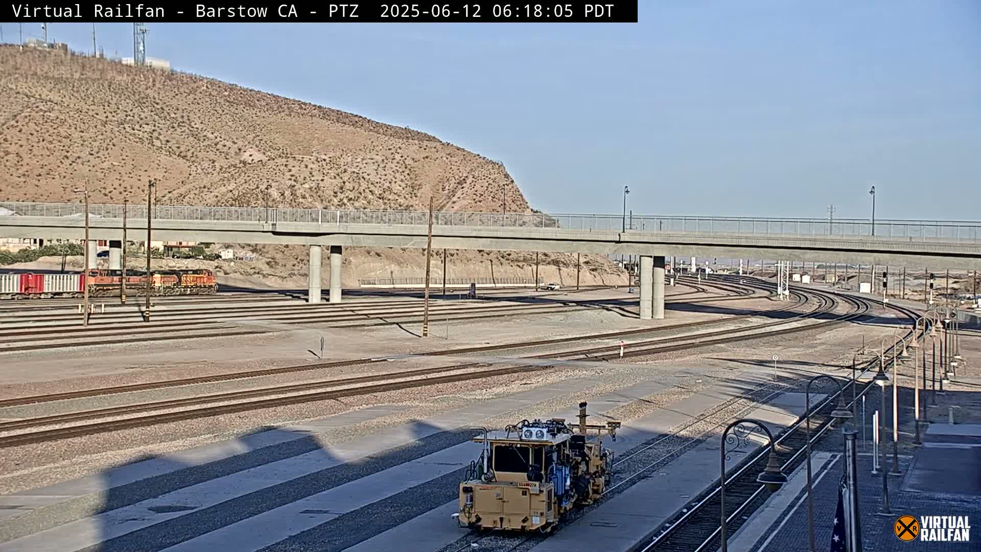 A sunny day reveals a railway yard with multiple tracks curving around a maintenance vehicle, all situated beneath a concrete overpass against a backdrop of a desert hillside.