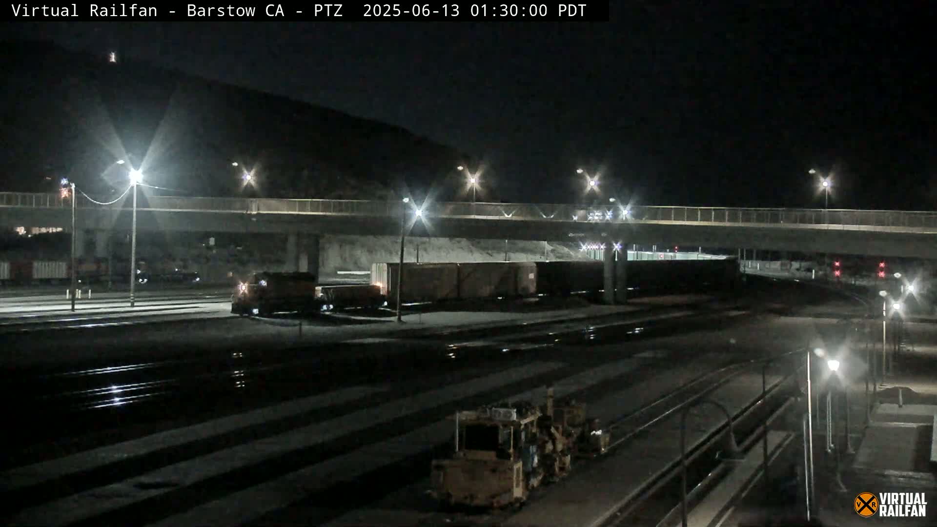 A nighttime view of a train yard with a train and other railway equipment under bright lights near a roadway overpass.