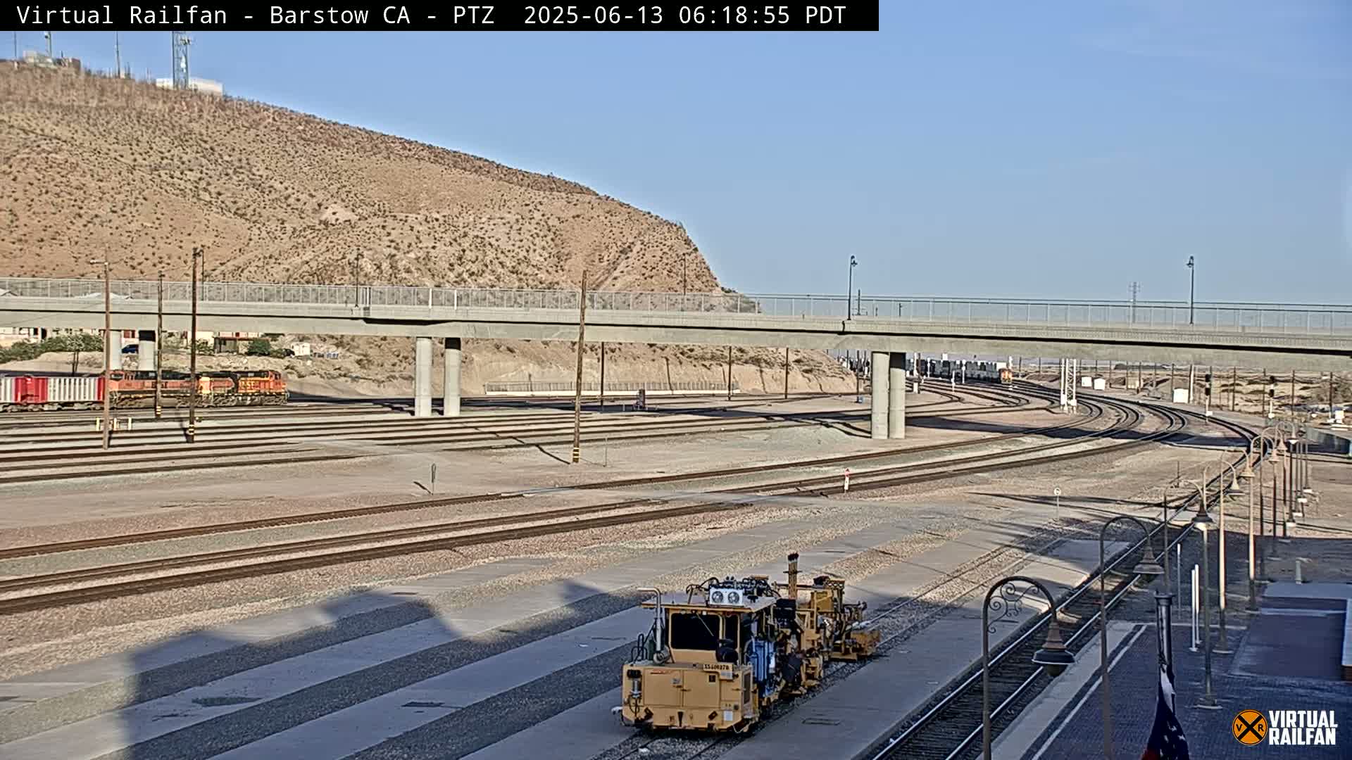 A rail yard with a train and a track maintenance vehicle, under a clear sky, next to a desert hillside and a bridge.