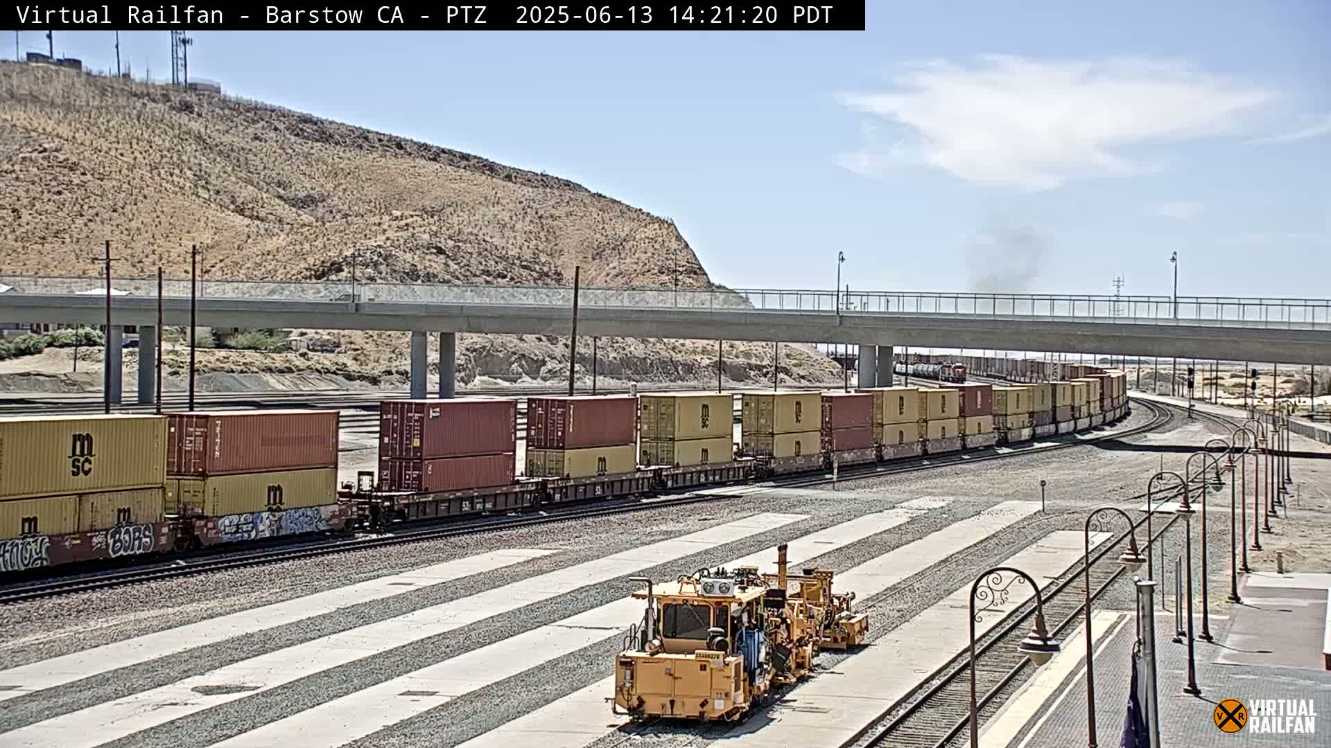 A long freight train carrying stacked shipping containers curves around a track under a highway overpass on a sunny day.