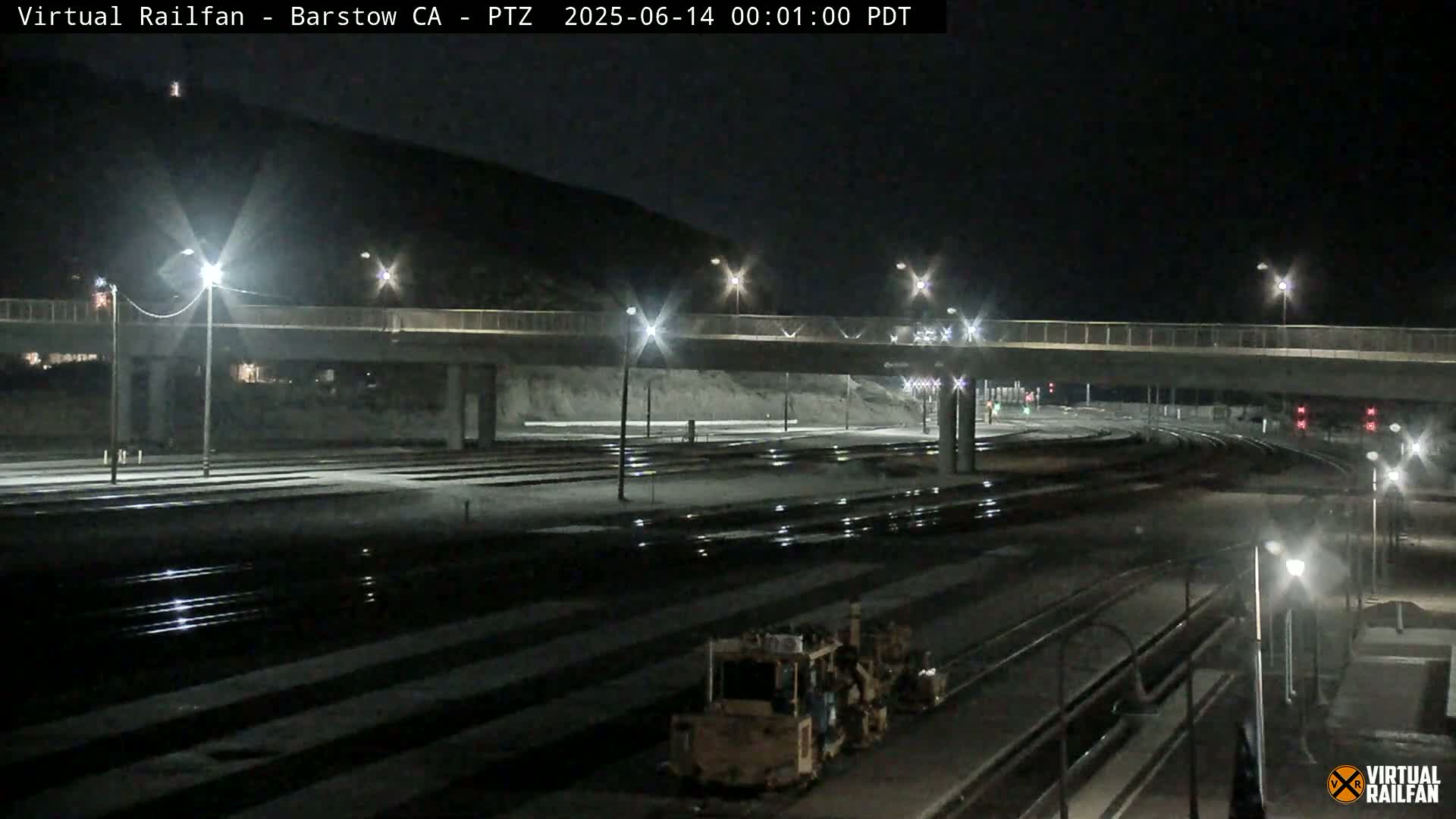 A nighttime view of a rail yard with a roadway overpass, illuminated by streetlights, showing a piece of construction equipment.