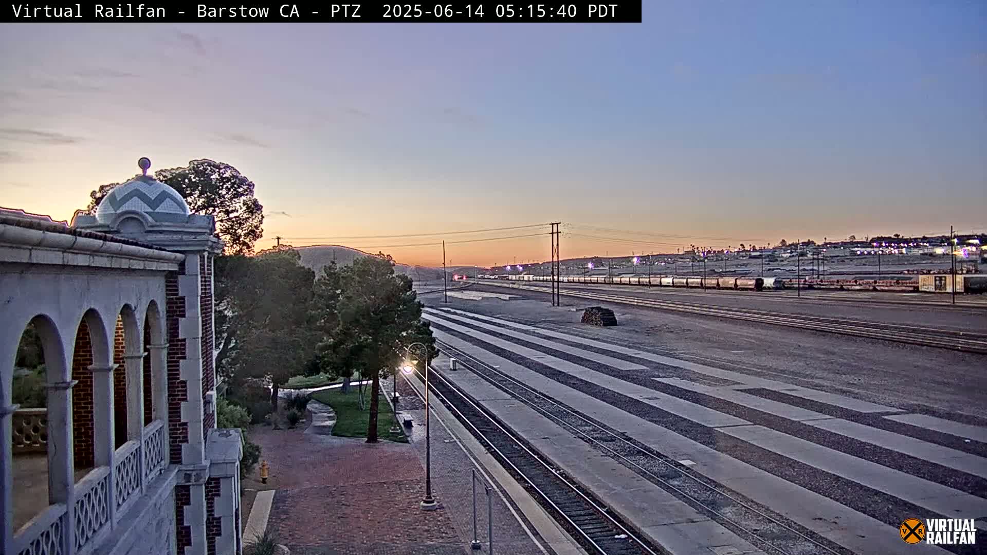 A long train sits on railway tracks at sunrise, viewed from an elevated position near a building with arched walkways.