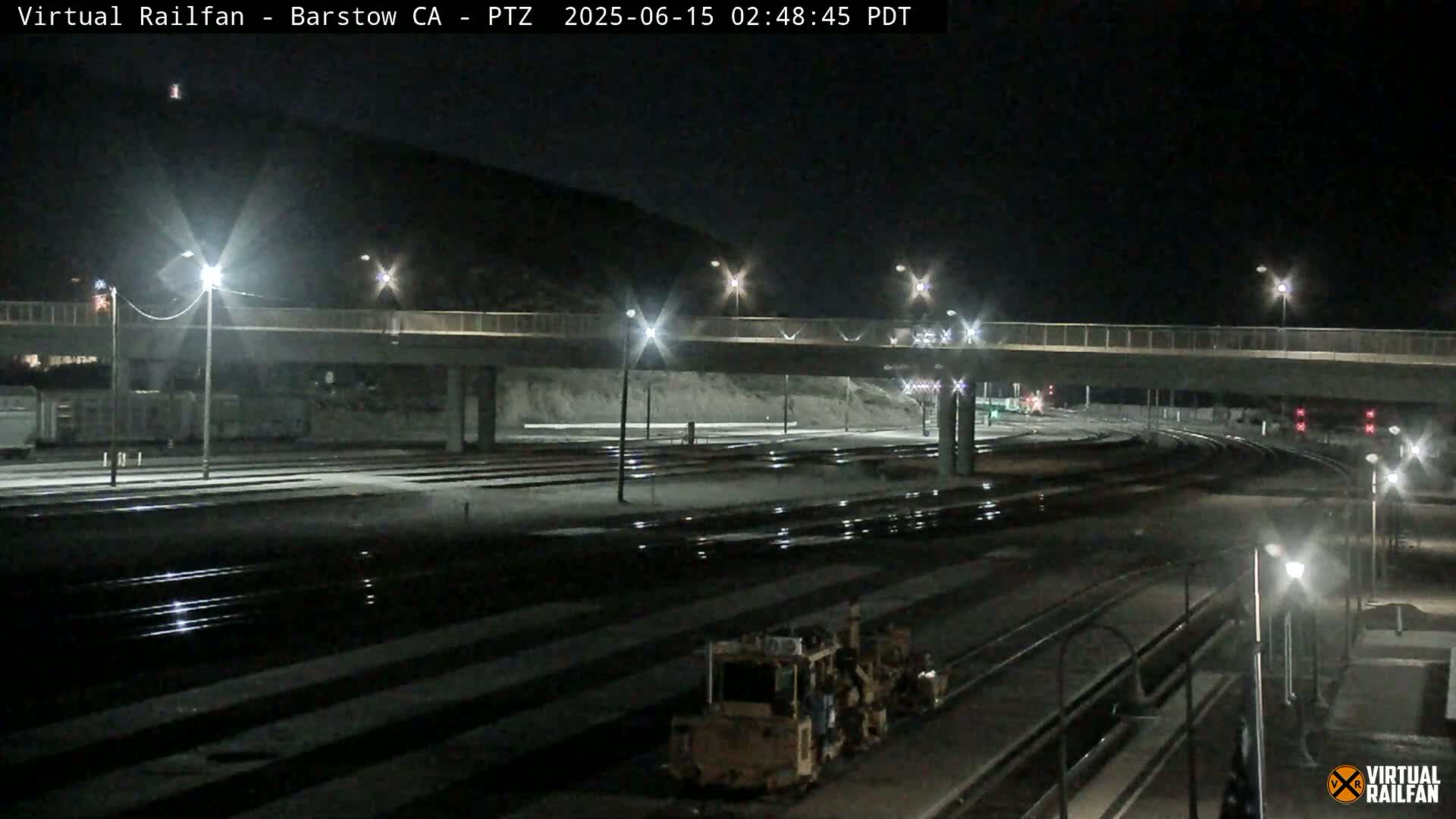 A nighttime view of a rail yard with a road overpass, illuminated by streetlights, showing train tracks and a piece of railway maintenance equipment.