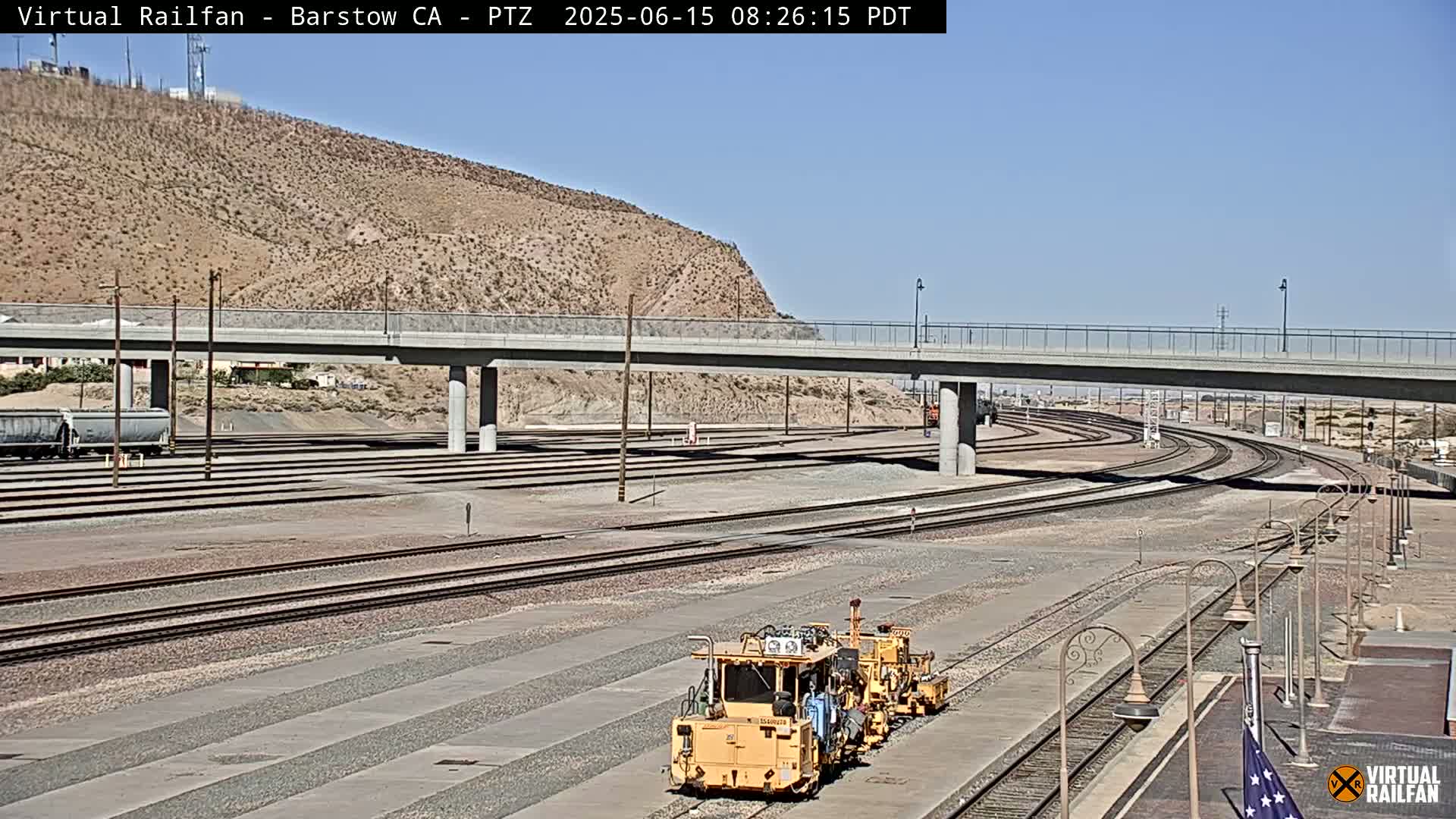 A yellow rail maintenance vehicle sits near a network of train tracks that extend under a bridge, all against a backdrop of a tan, arid hill under a clear sky.