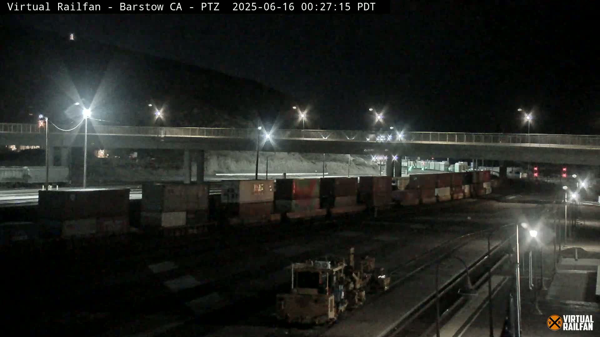 At night, a train carrying multiple shipping containers sits below a highway overpass, illuminated by streetlights.