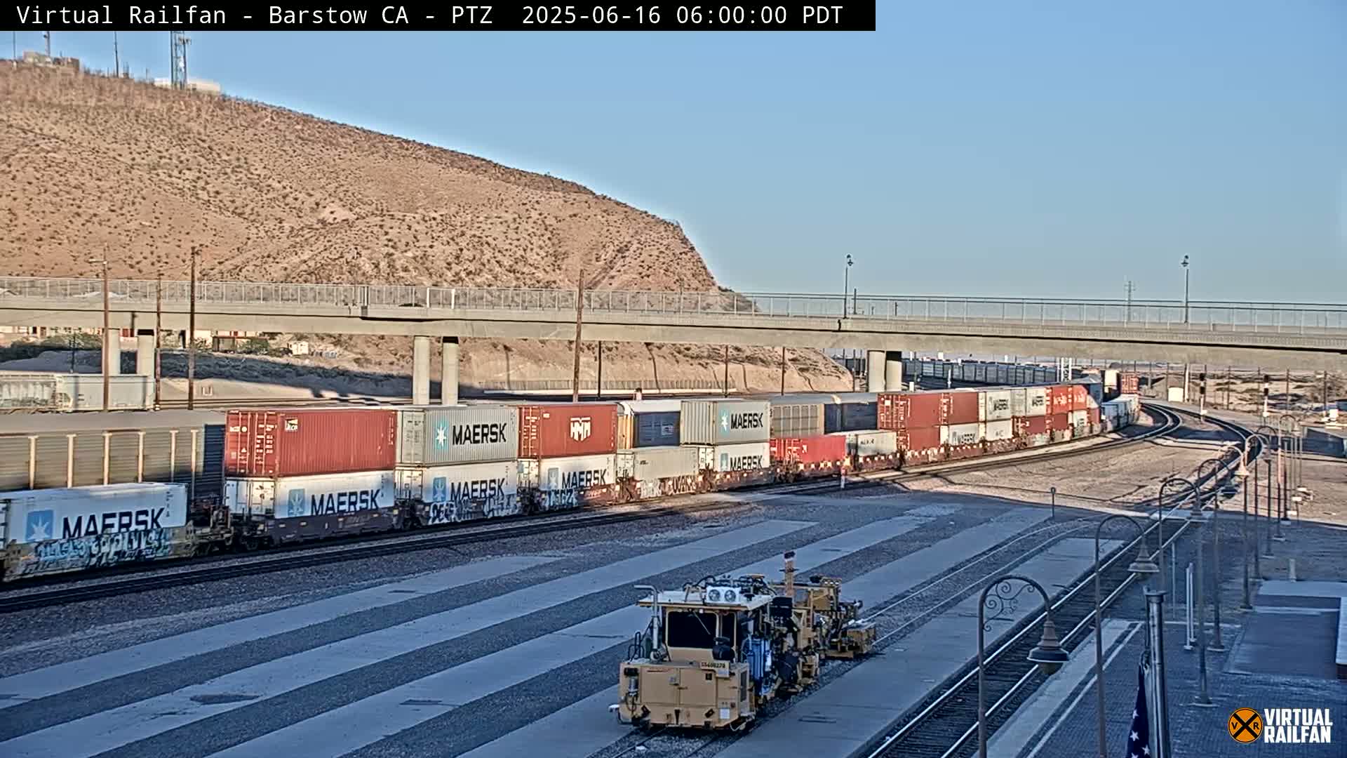 Under a clear sky, a long freight train carrying numerous shipping containers sits on the tracks next to a rail maintenance vehicle near a highway overpass and a desert hillside.