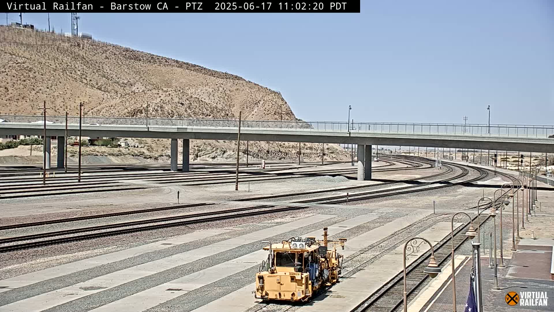 A yellow rail maintenance vehicle sits in a rail yard under a sunny sky, with a road overpass visible in the background.