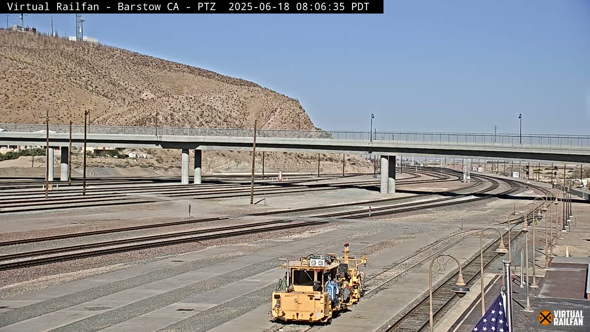 A yellow rail maintenance vehicle sits on a network of train tracks under a bridge on a sunny day.