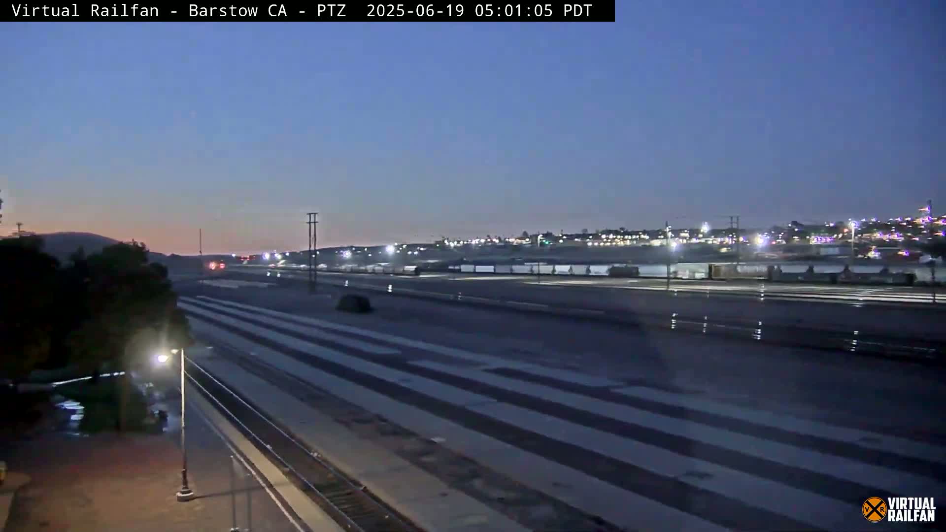 Under a twilight sky, a wide shot shows a train yard with multiple parallel tracks and a distant cityscape at dawn.