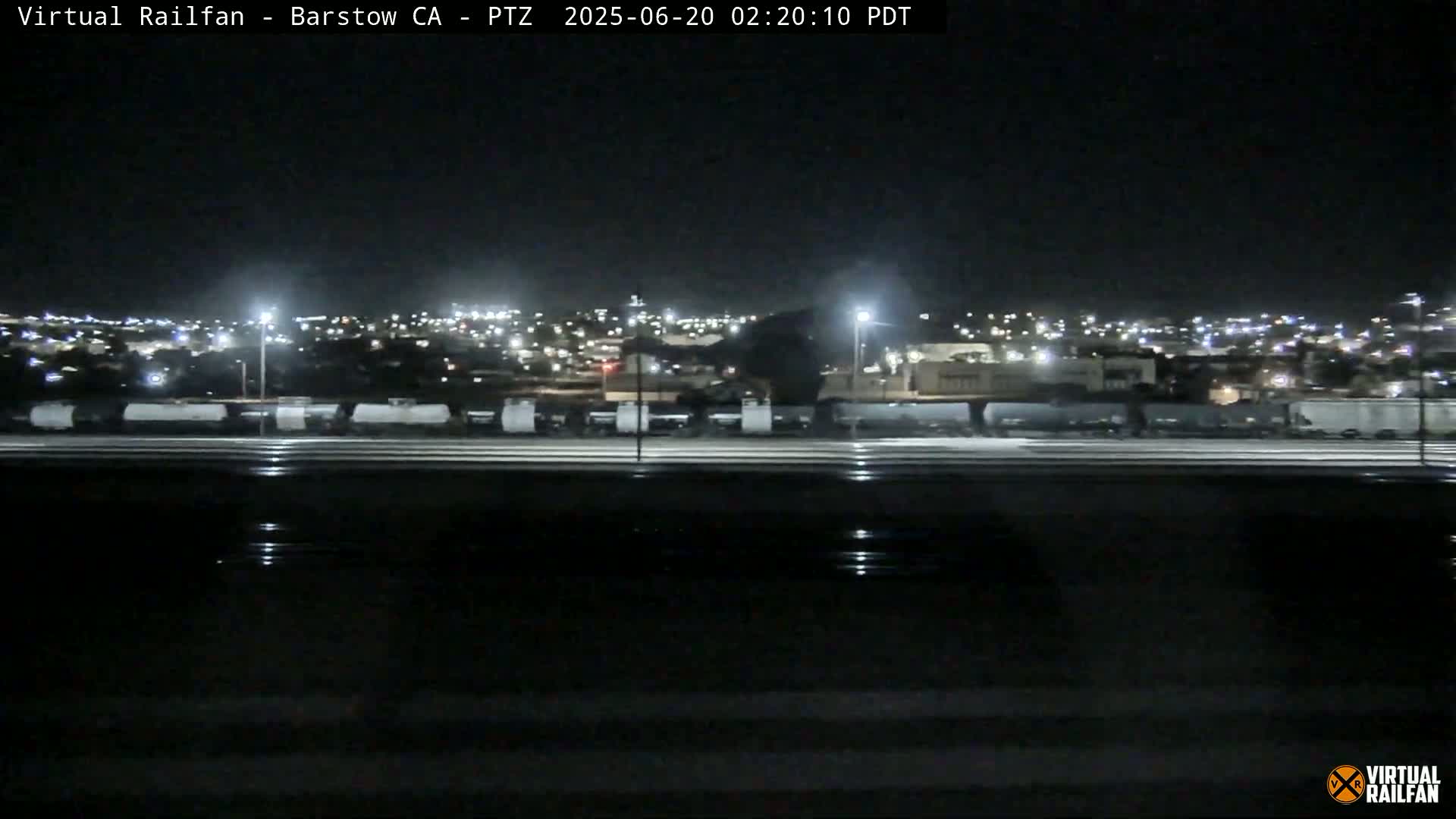 A nighttime view of a train yard and city lights beyond, with a dark, possibly wet road in the foreground.