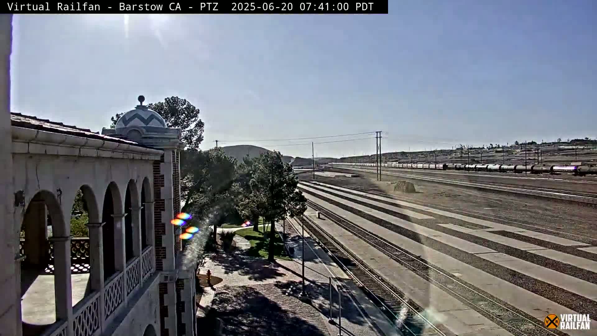 A long freight train is visible on a sunny day, passing by a section of railway tracks next to a building with arched walkways and a domed roof.