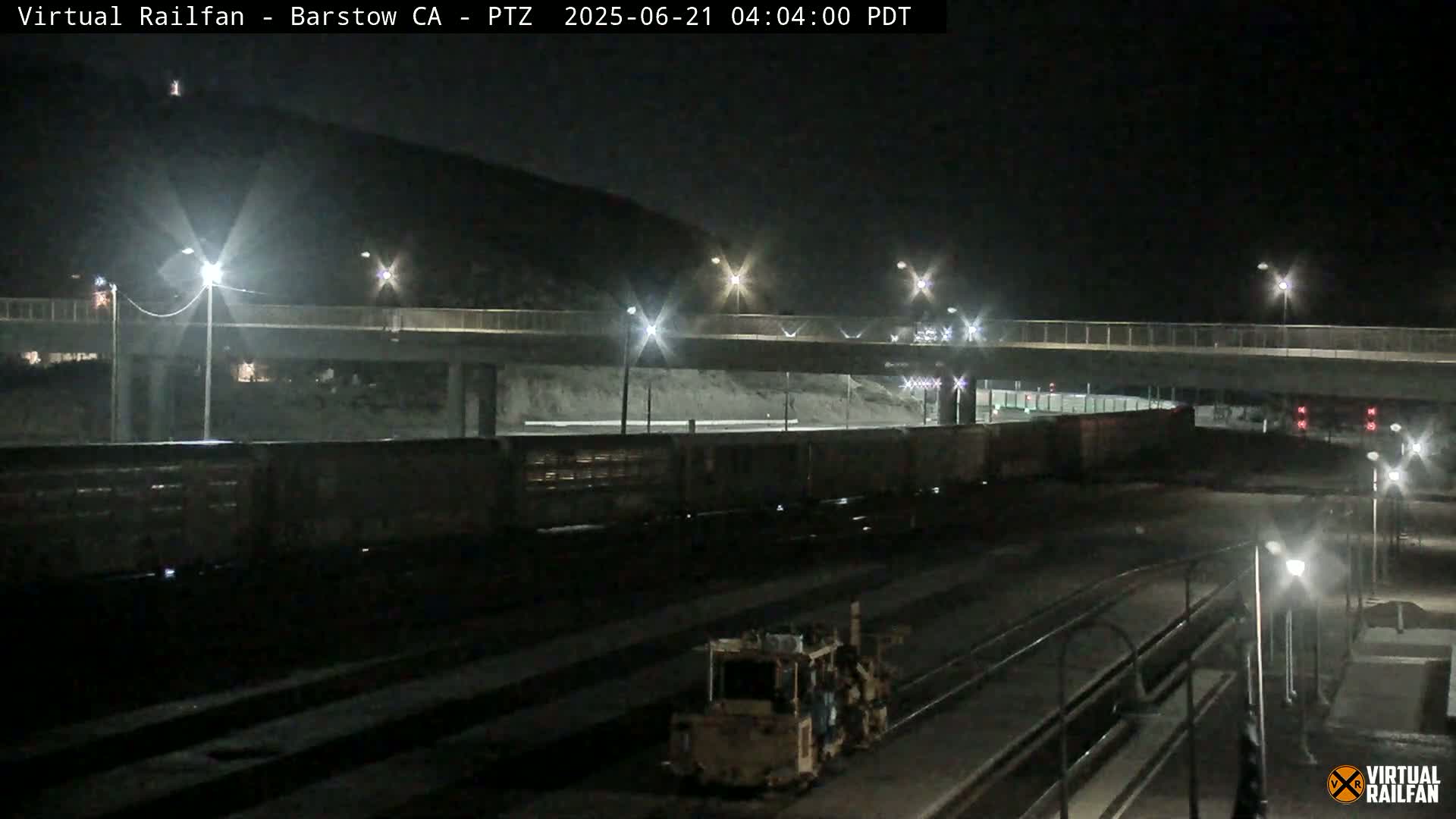 A long freight train sits under a highway overpass at night, with a piece of railway maintenance equipment visible in the foreground.