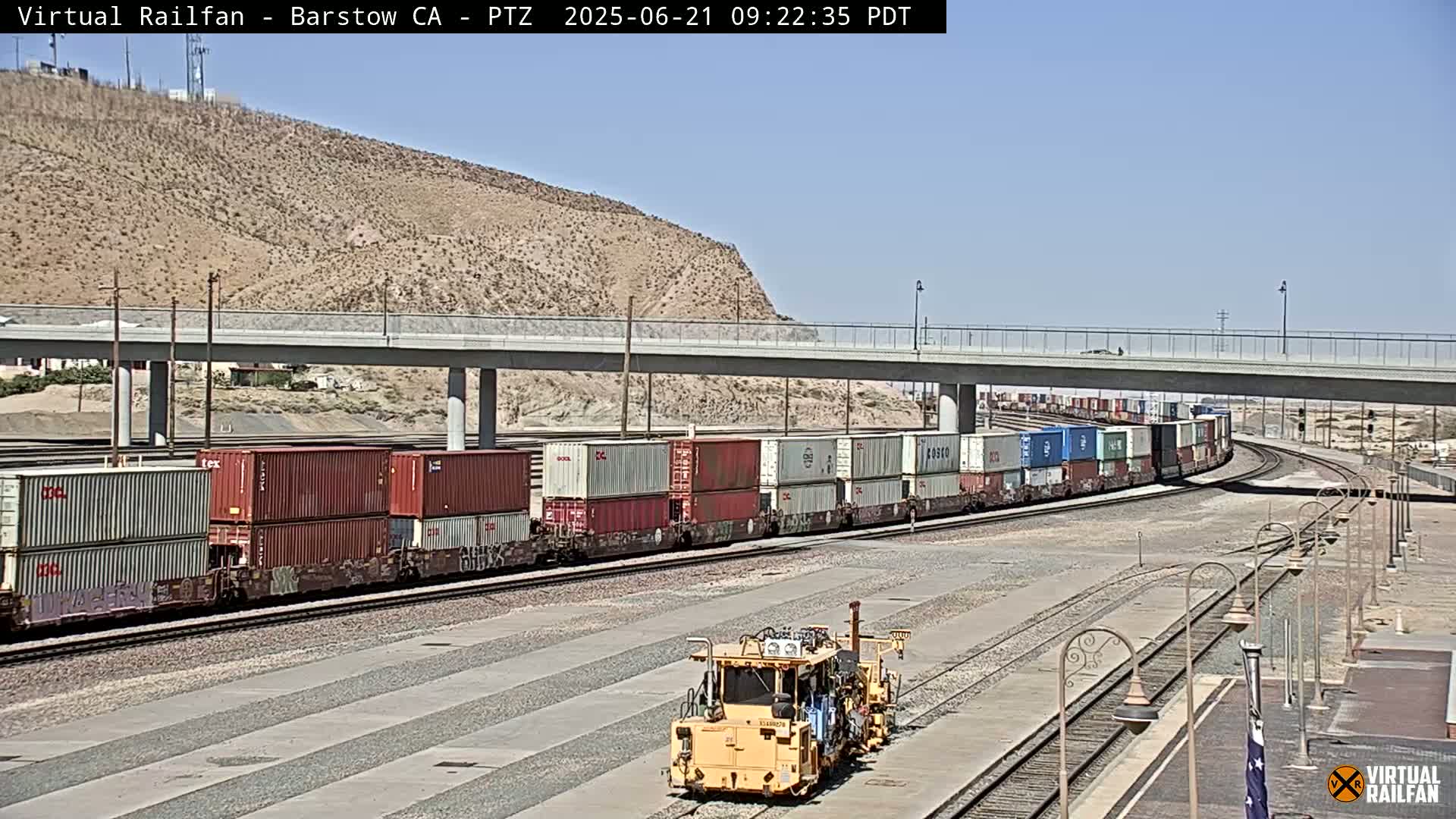 A long freight train carrying multiple shipping containers passes under a bridge on a sunny day in a desert landscape.