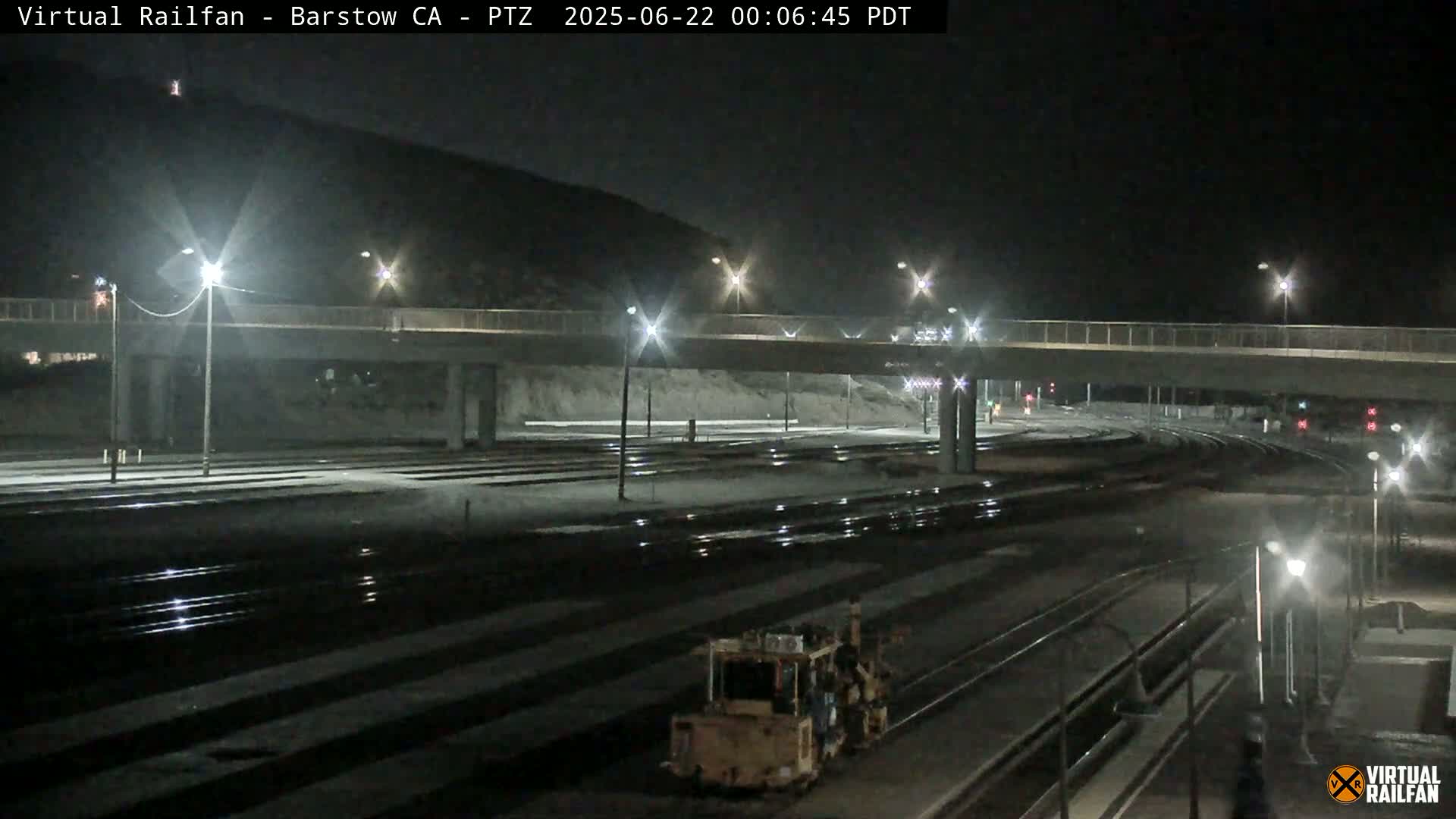 A nighttime view of railroad tracks and a roadway overpass, illuminated by streetlights, with a piece of railway maintenance equipment visible in the foreground.