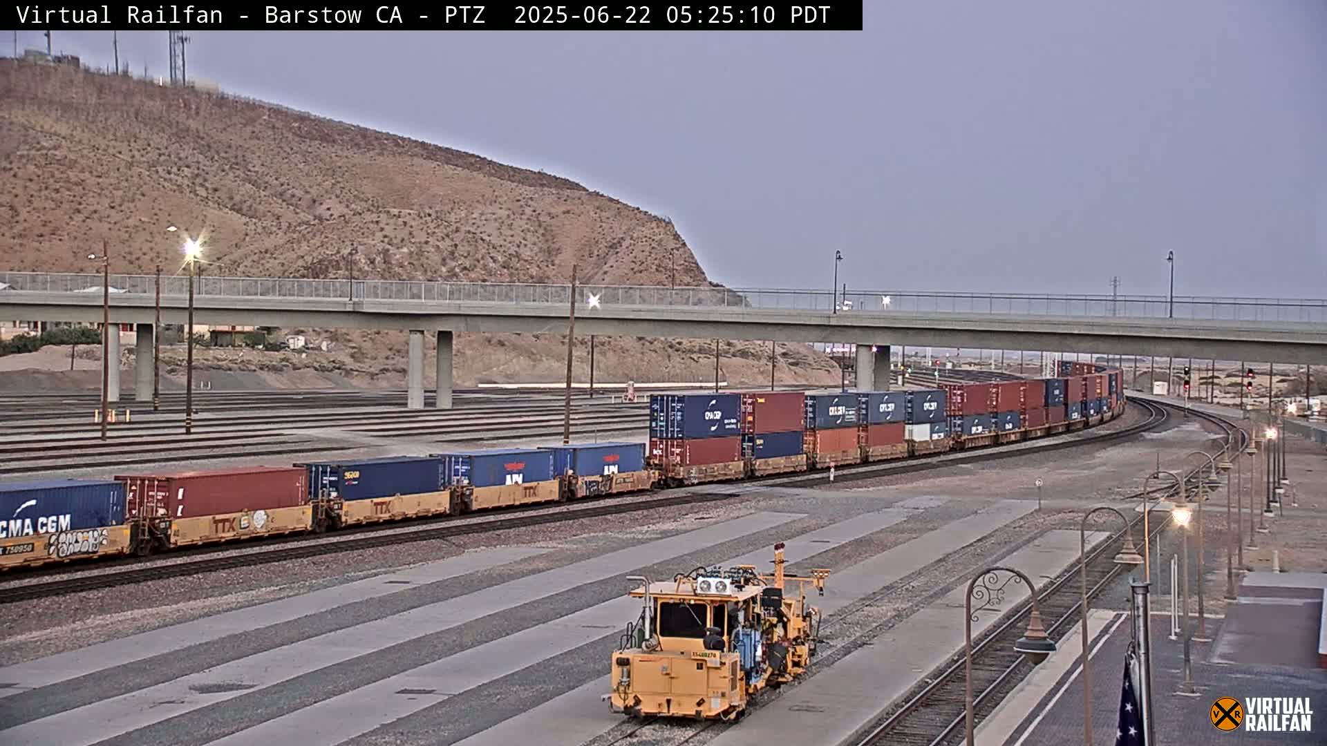 A long freight train carrying multiple shipping containers curves around a track under a highway overpass in the early morning twilight.