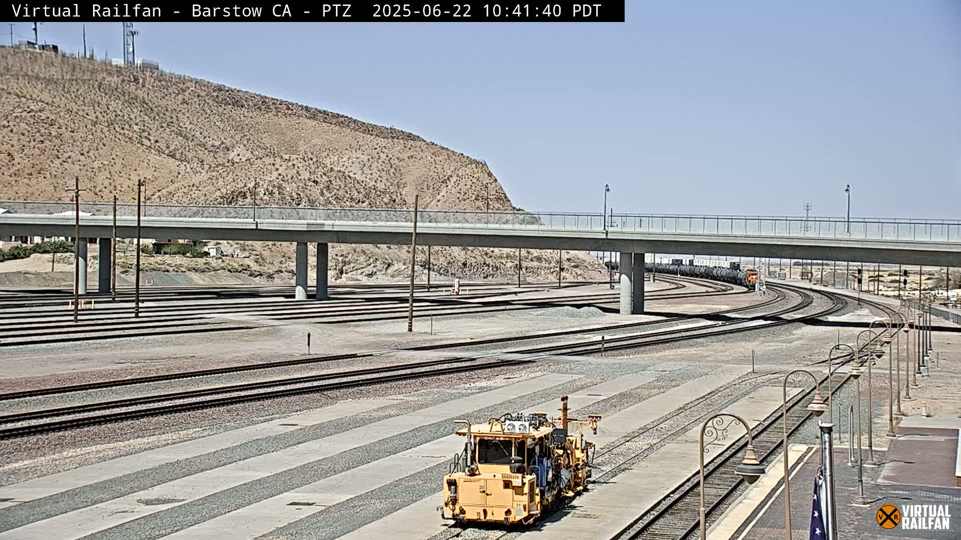 A yellow rail maintenance vehicle sits in a rail yard under a bridge on a sunny day.