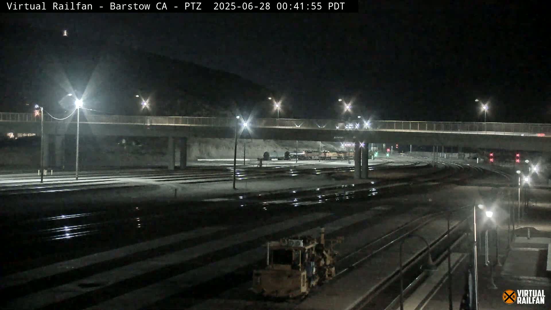 A nighttime view of a rail yard with a road overpass, lit by streetlights, showing some railway maintenance equipment.