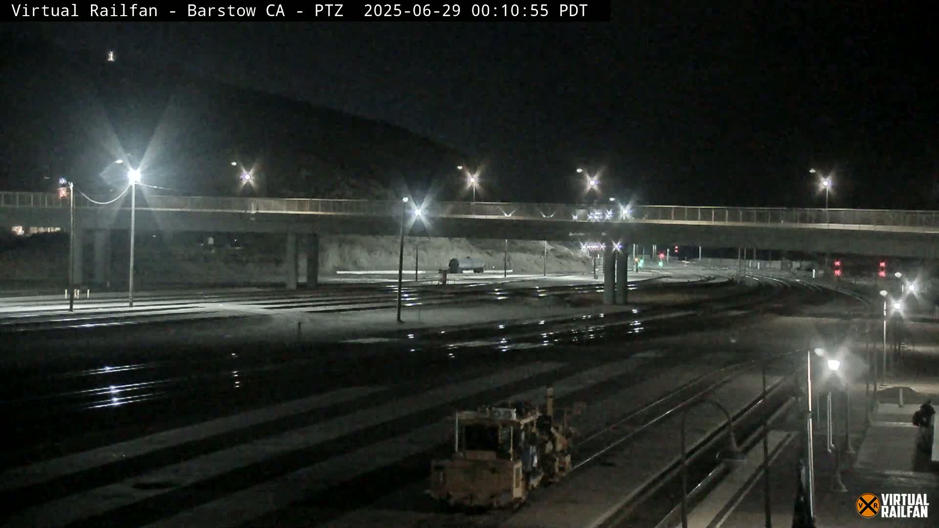 A nighttime view of a railway yard with a road overpass, illuminated by streetlights, under a clear, dark sky.