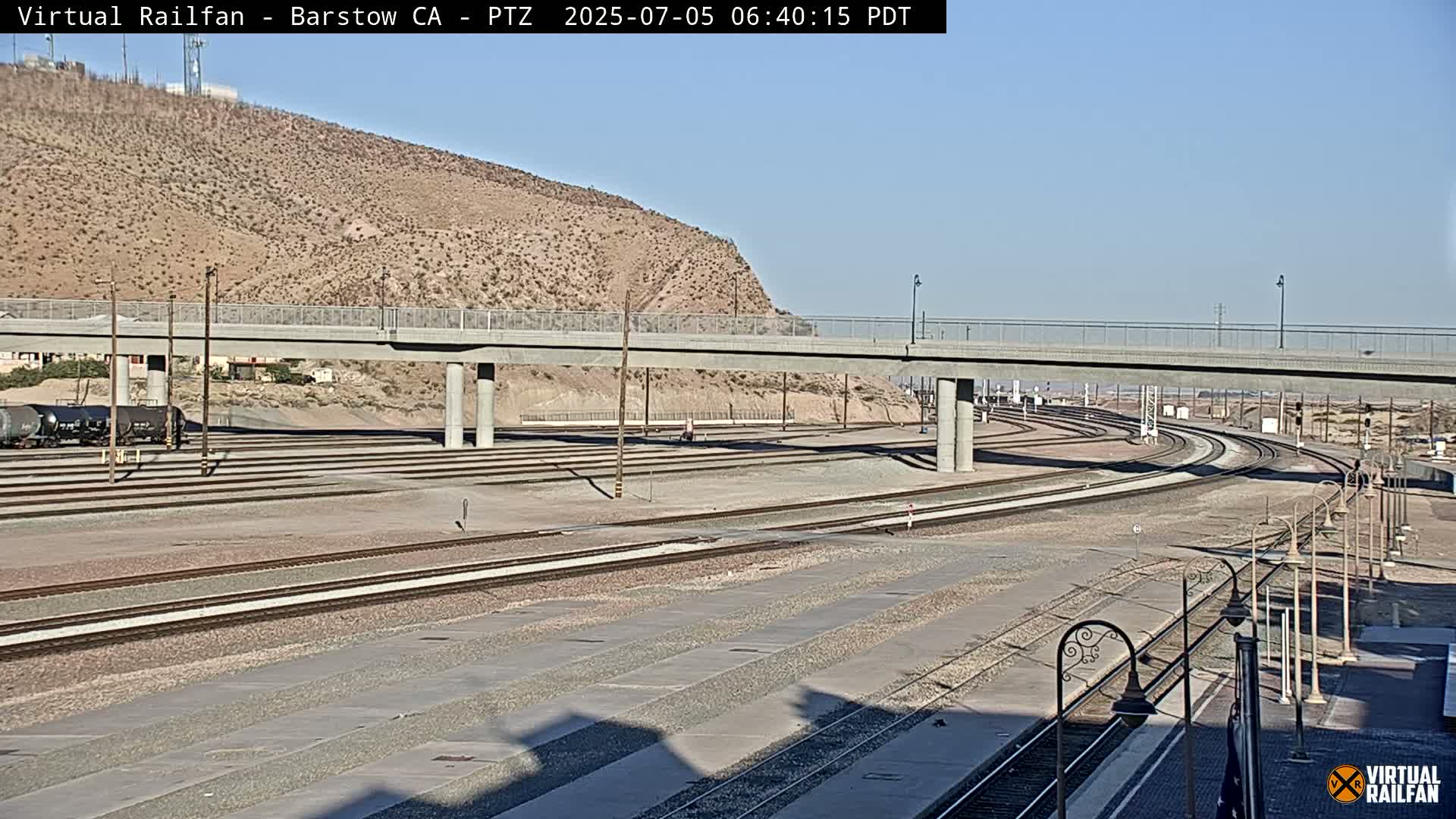 A railway yard with multiple tracks under a bridge on a clear, sunny day, set against a backdrop of a tan desert mountain.
