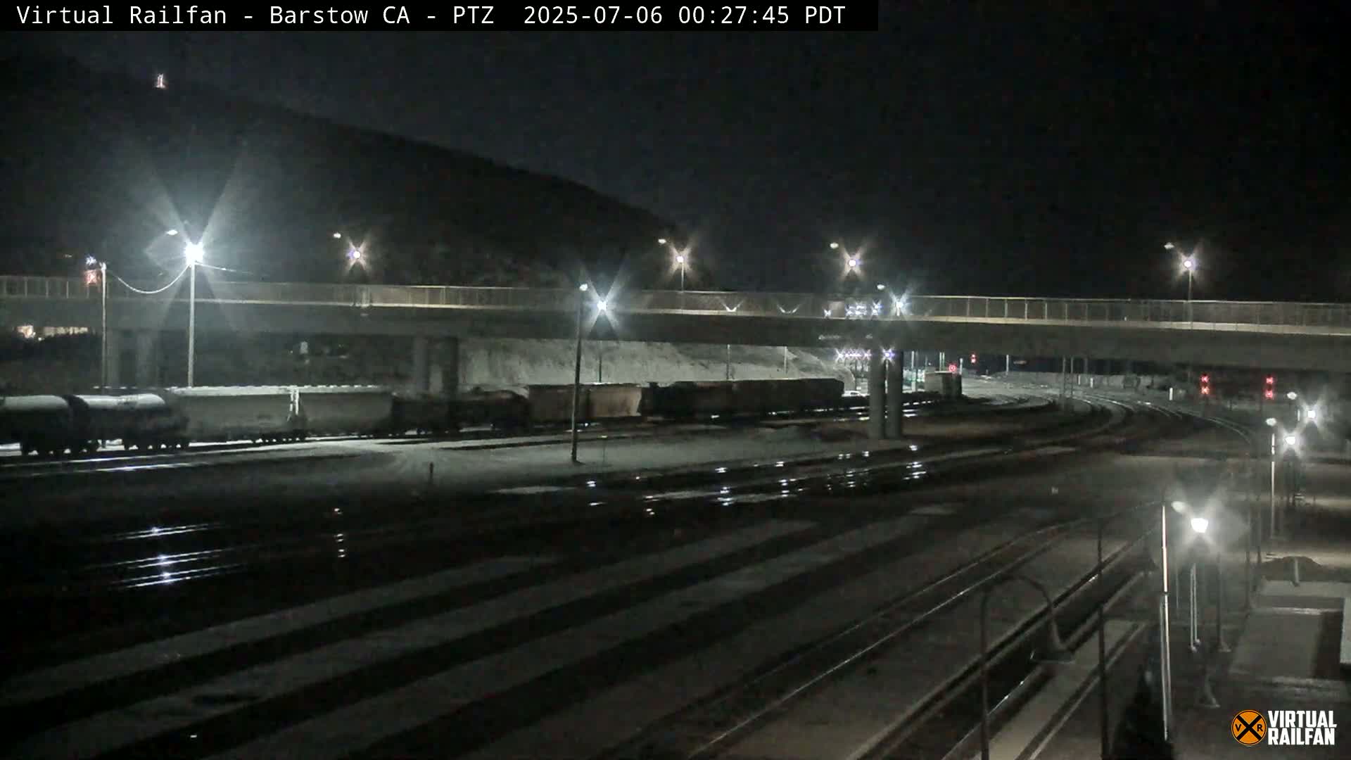 A nighttime view of a rail yard with several train cars parked under a bridge, lightly snow-covered ground, and several bright streetlights.