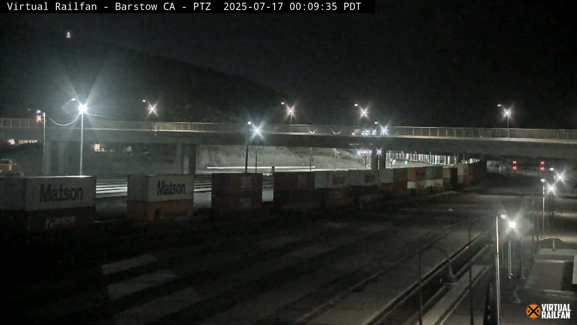 At night, under clear skies, a line of freight cars sits beneath a highway overpass.