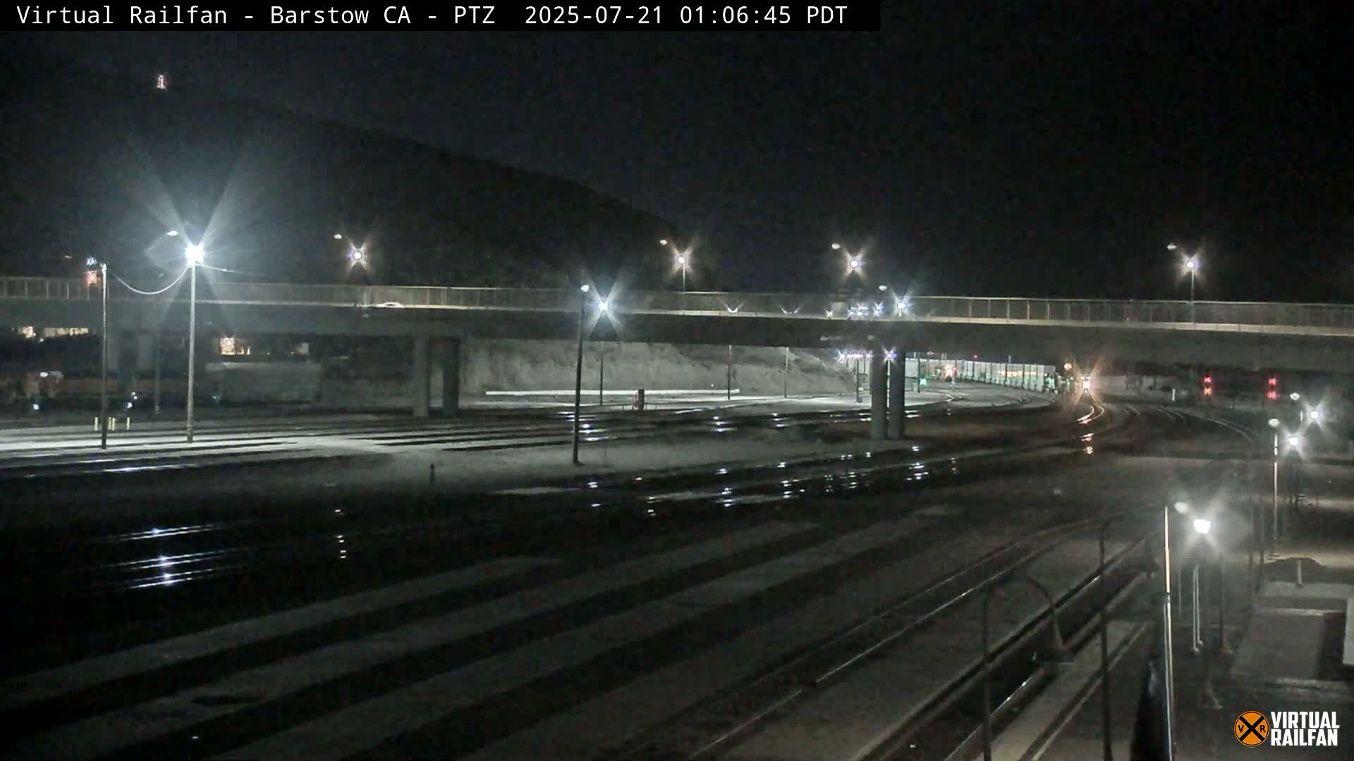 A nighttime view of a train yard with multiple sets of train tracks, a roadway overpass, and several bright lights illuminating the scene.