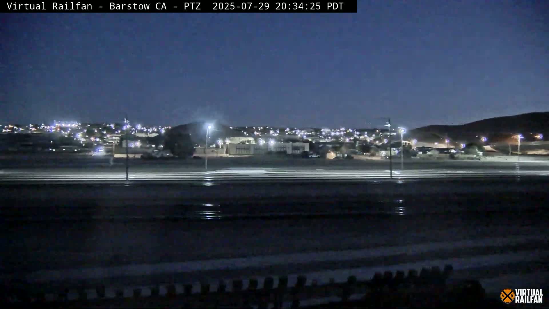 A nighttime view of a flat, dark expanse of land with parallel lines, a small piece of machinery, and distant lights under a clear, dark sky.