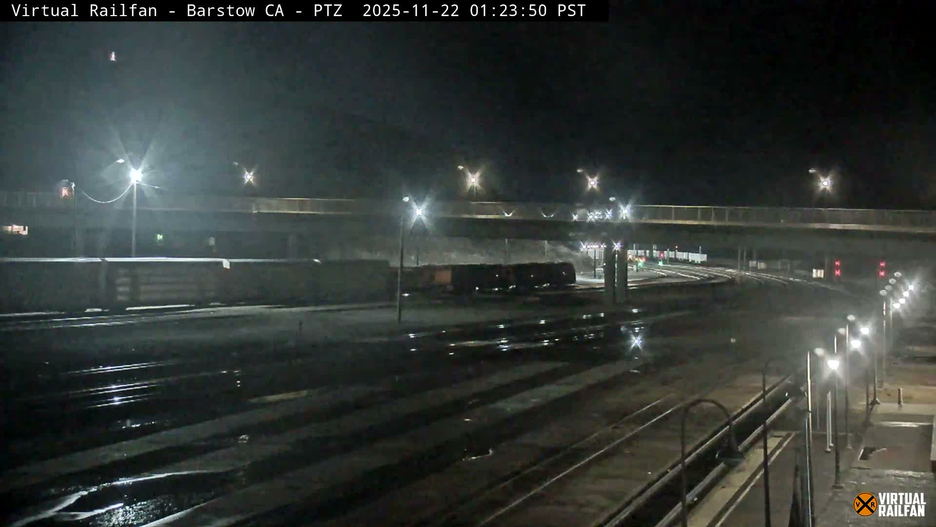 A nighttime view of a wet railyard features numerous tracks reflecting bright streetlights under an overhead bridge, with train cars visible in the distance.