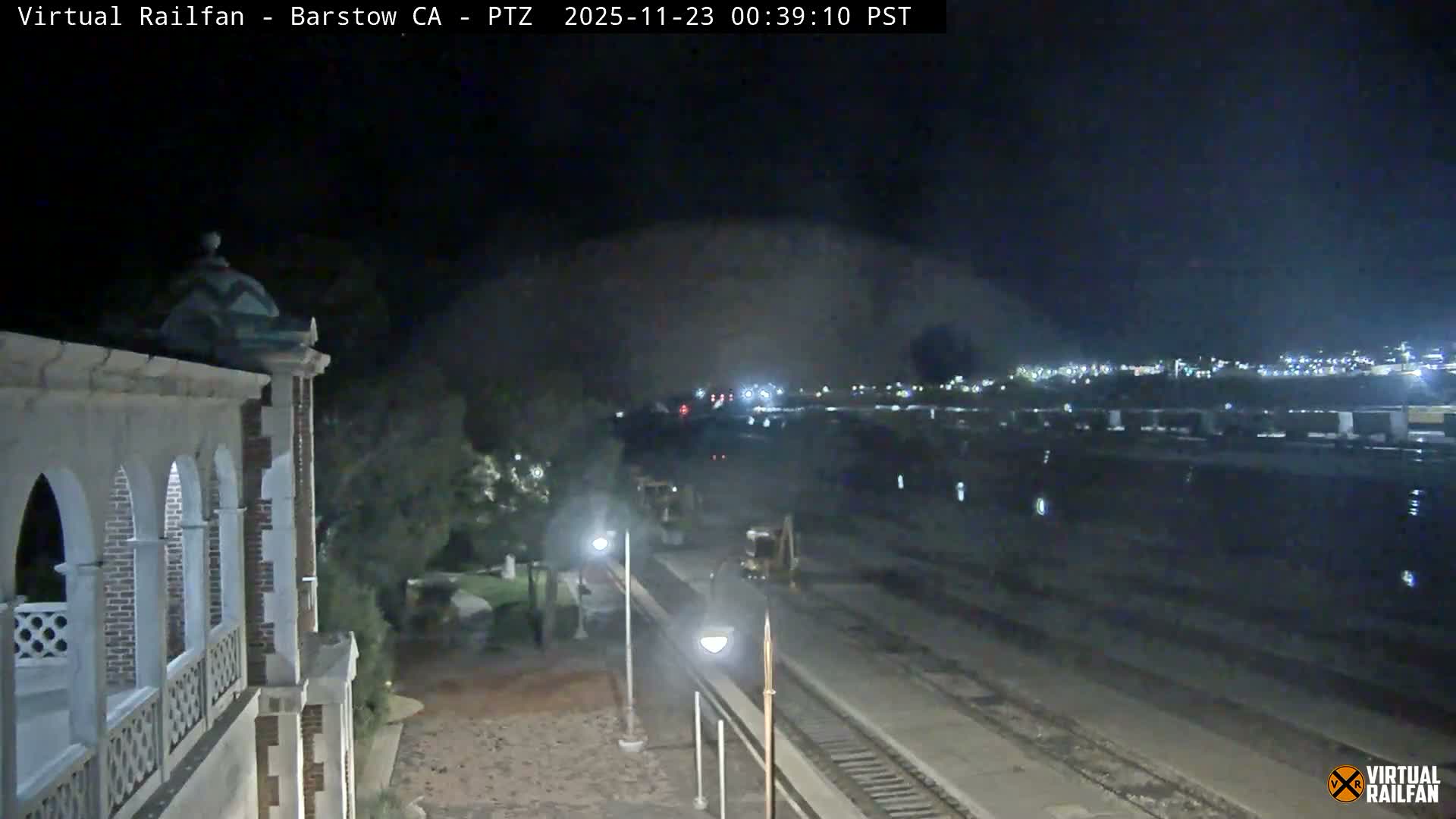 An ornate arched building stands beside multiple railway tracks at night, where a train with a bright headlight and several construction vehicles are visible, with a distant city glowing under a clear sky.