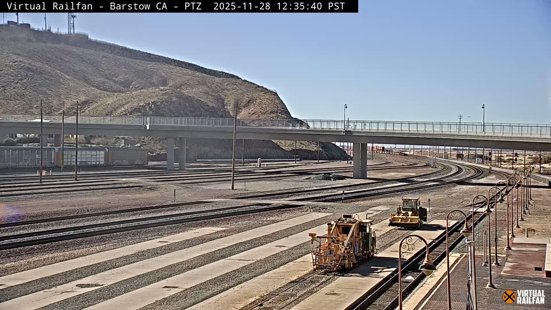 The image captures a sunny outdoor scene of a large railway yard with multiple tracks, featuring two yellow rail maintenance machines, a concrete overpass, and a barren brown hill under a clear blue sky.