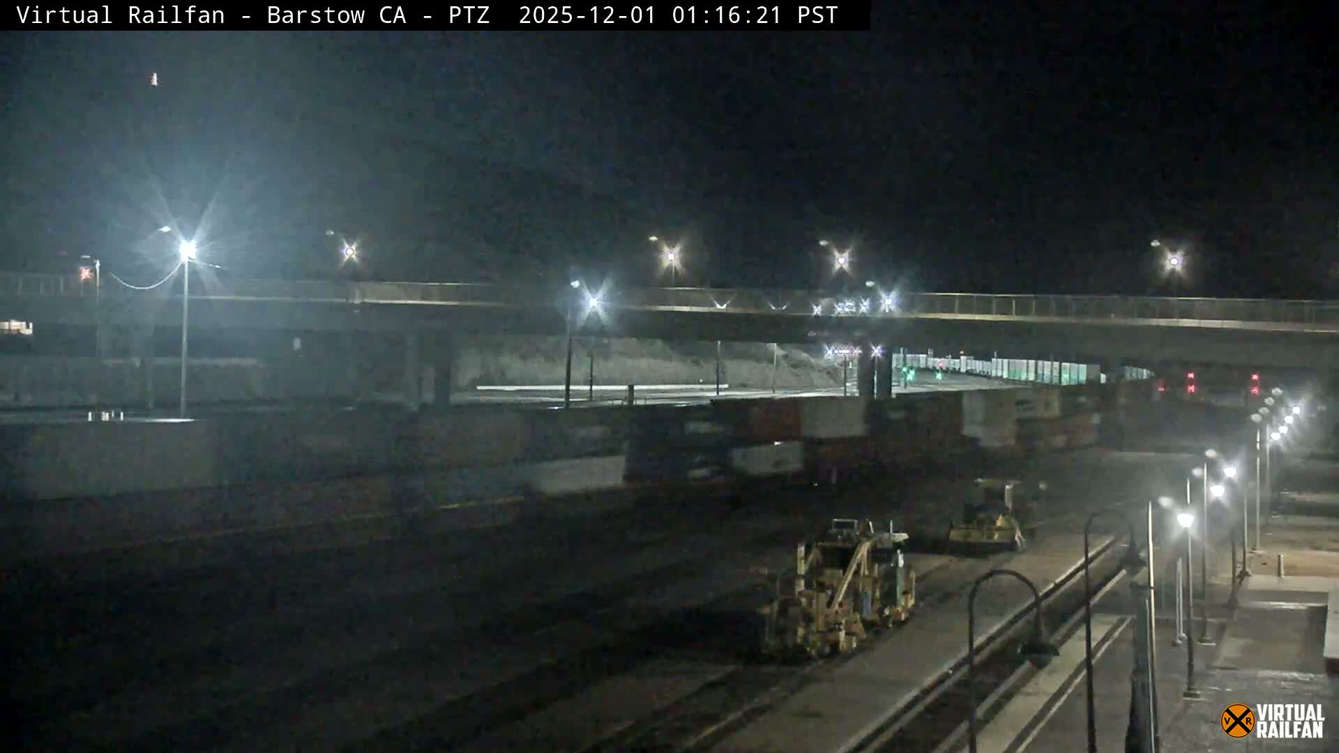 The image shows a clear, dark night scene at a rail yard featuring multiple train tracks, several rail maintenance vehicles, an overpass, and numerous shipping containers in the background.
