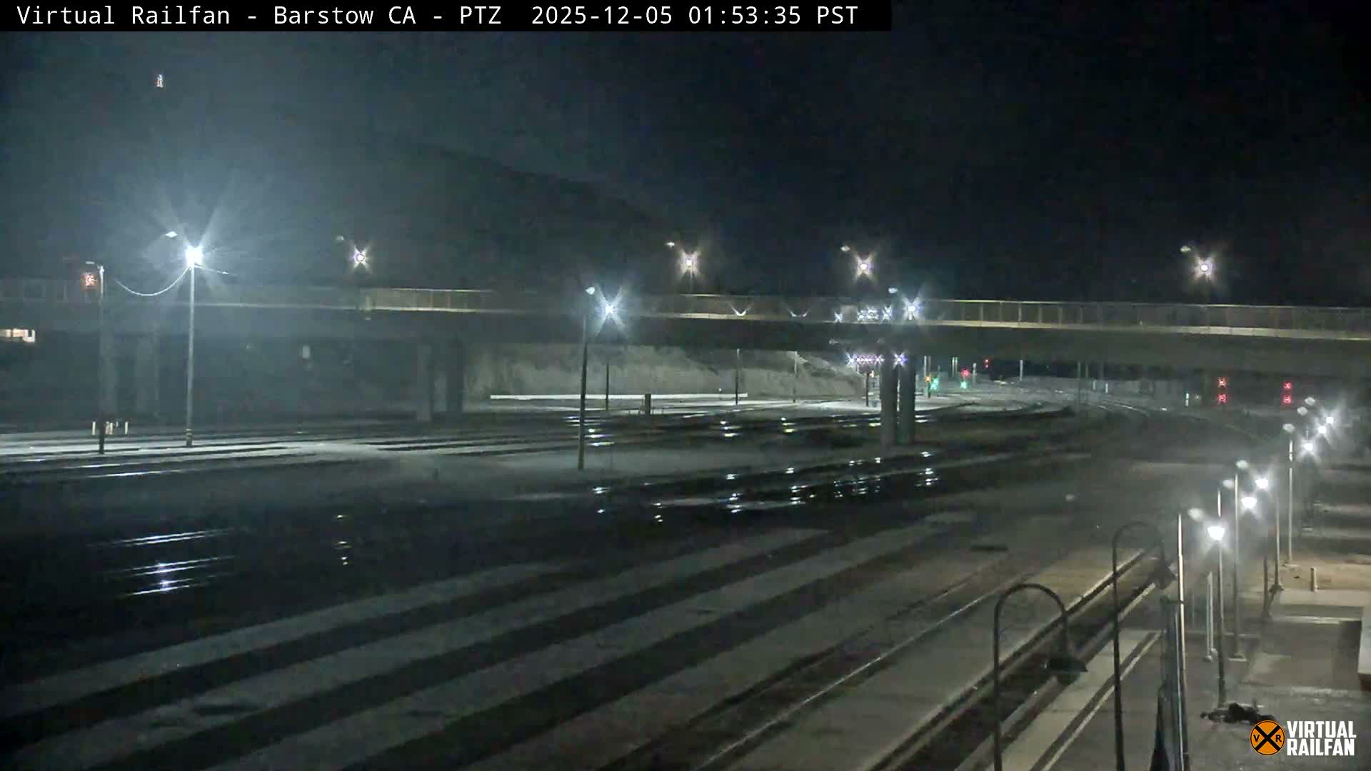 A wide view of a train yard at night, illuminated by numerous streetlights reflecting off damp surfaces, features multiple tracks, railway signals, and a concrete overpass against a dark, clear sky.