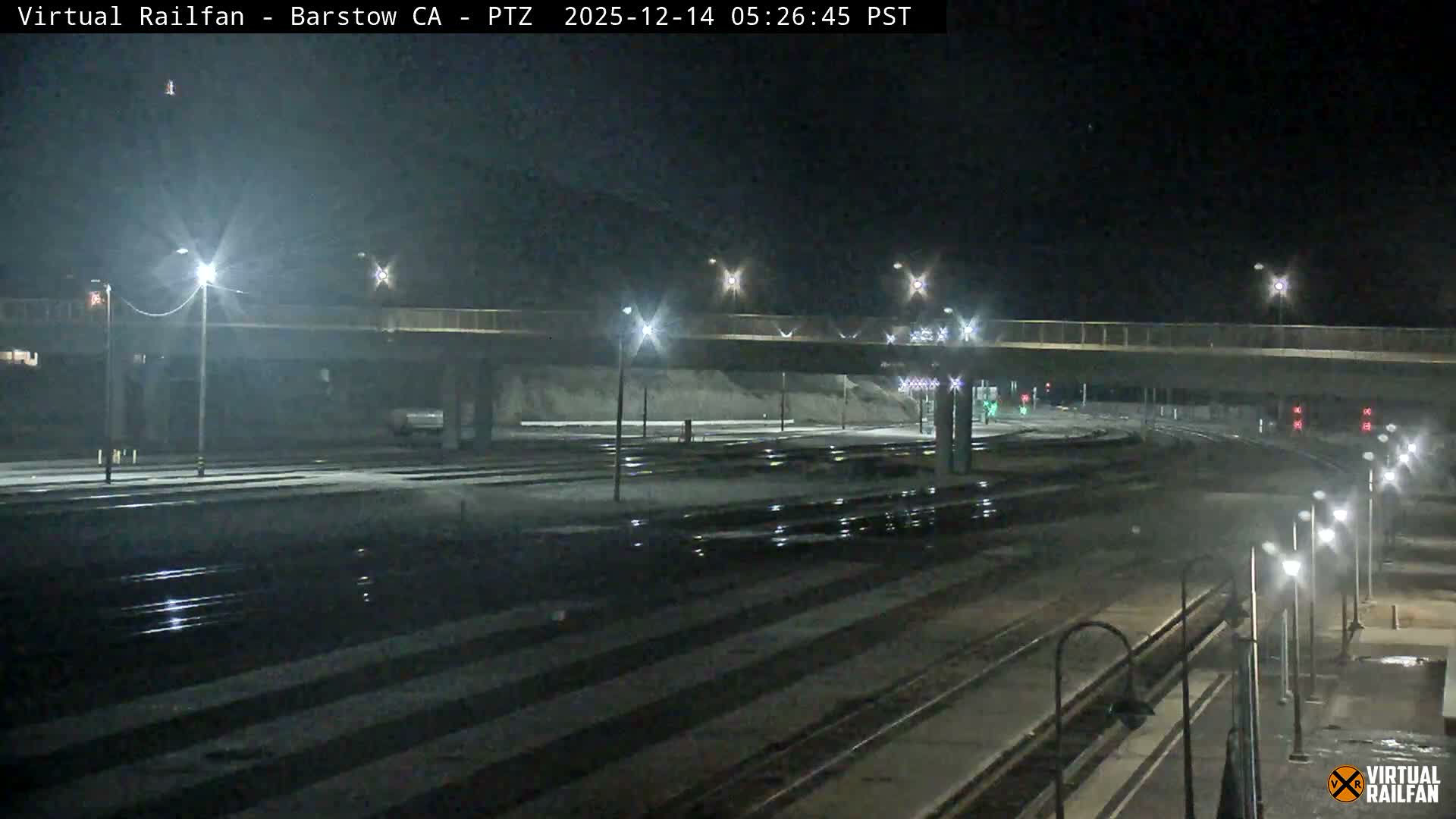 A wide view of a train yard at night, illuminated by numerous streetlights reflecting off damp surfaces, features multiple tracks, railway signals, and a concrete overpass against a dark, clear sky.