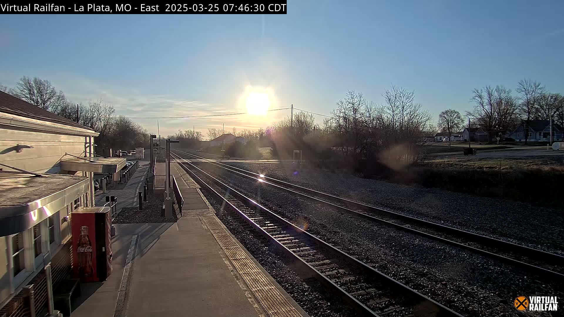 A sunny morning view of a train station platform with two sets of train tracks extending into the distance, alongside a residential area.