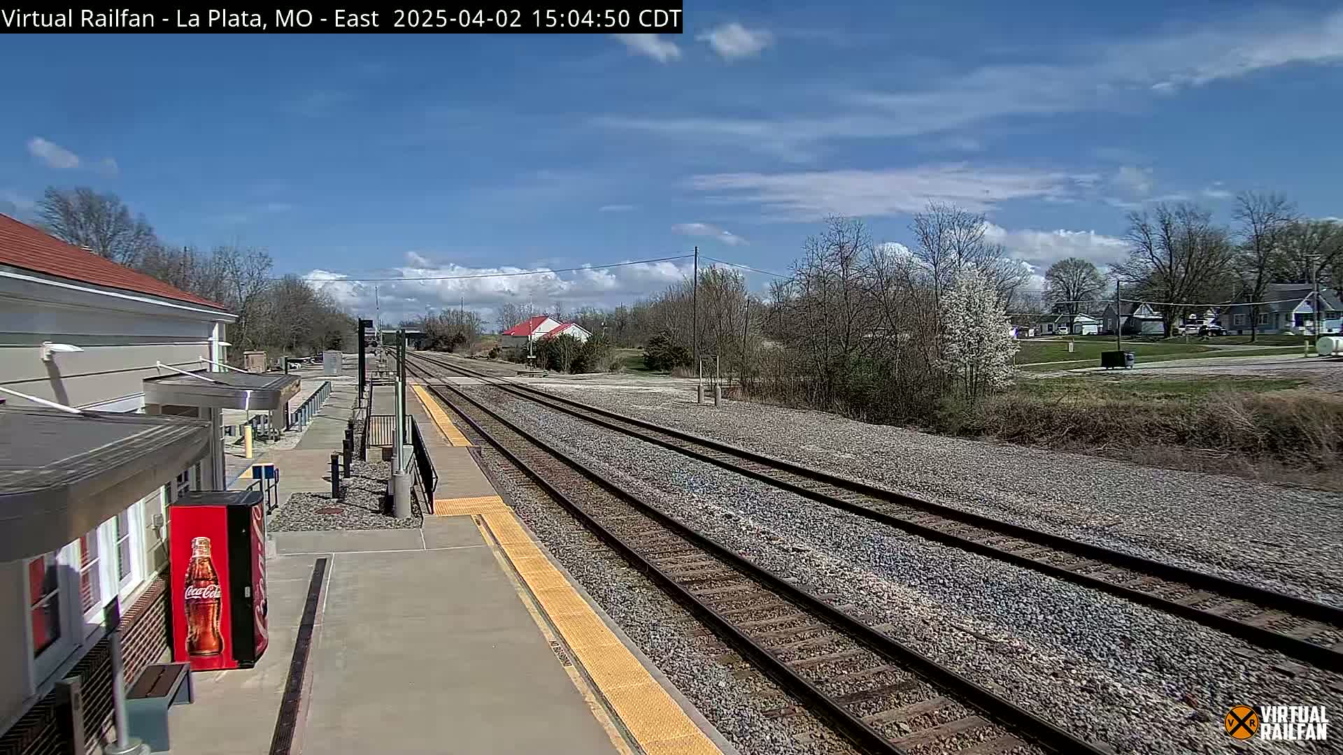 A mostly sunny day shows a train station platform with tracks extending into the distance,  surrounded by trees and houses.