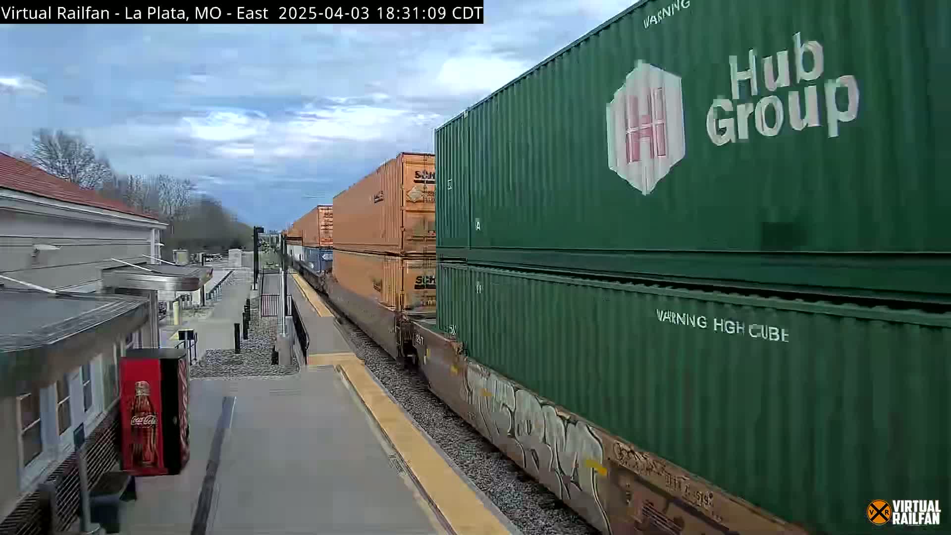 A long freight train carrying orange and green shipping containers passes a train station on a partly cloudy day.