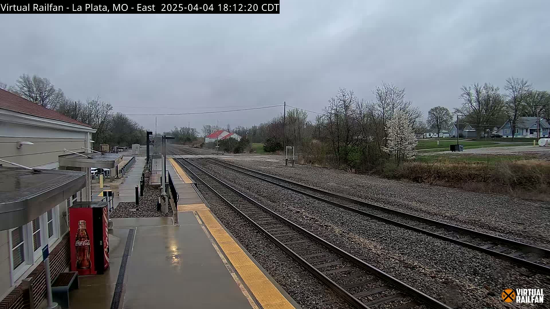 A wet train platform and tracks extend into a landscape of houses and trees under an overcast sky.