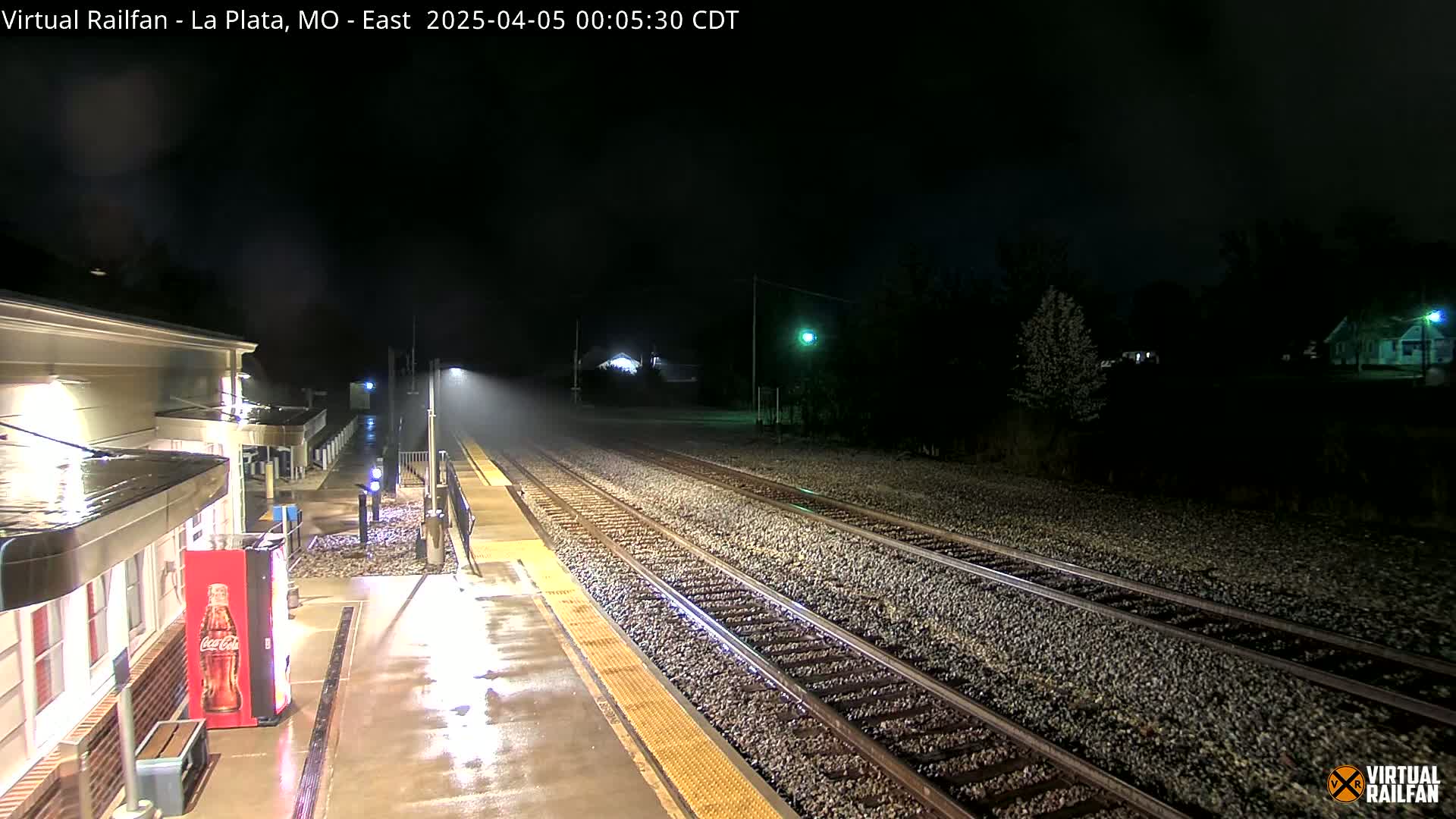 A nighttime view of a rain-slicked train station platform and tracks, illuminated by streetlights.