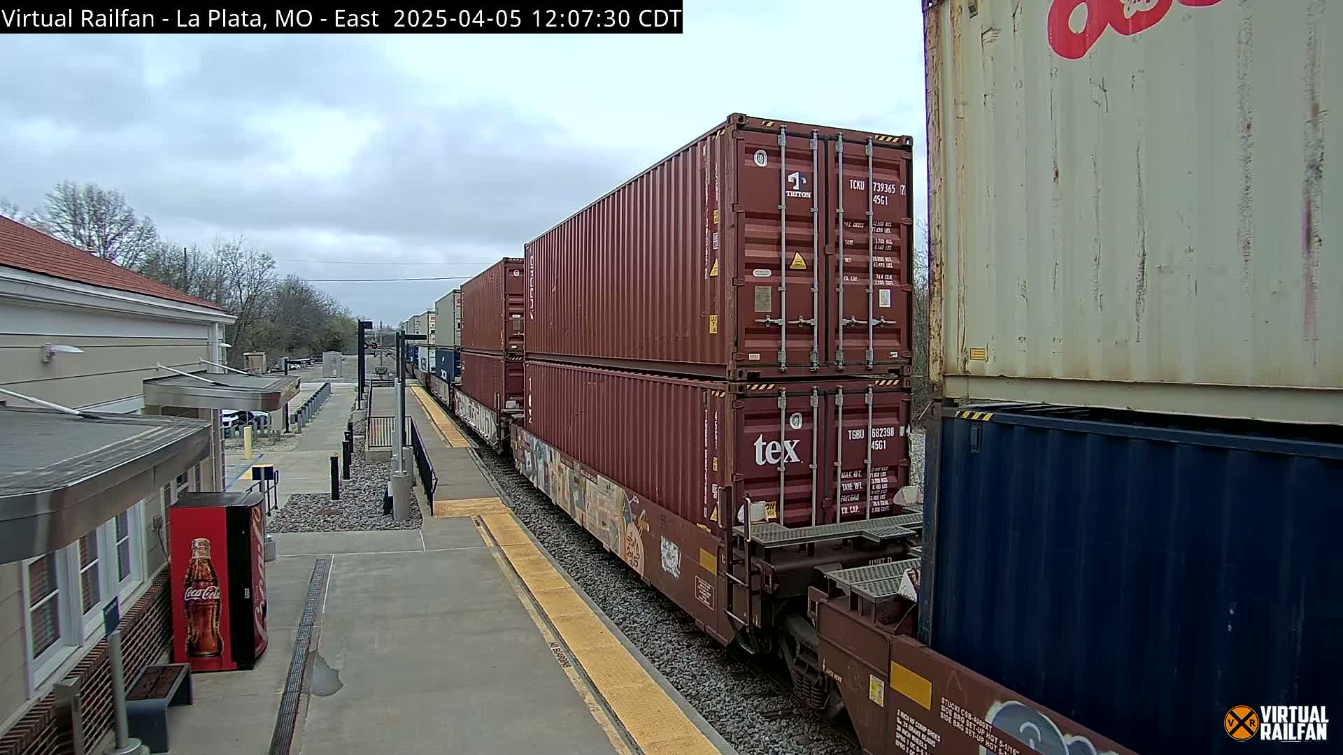 A freight train carrying stacked shipping containers is seen on a track next to a station building under a cloudy sky.