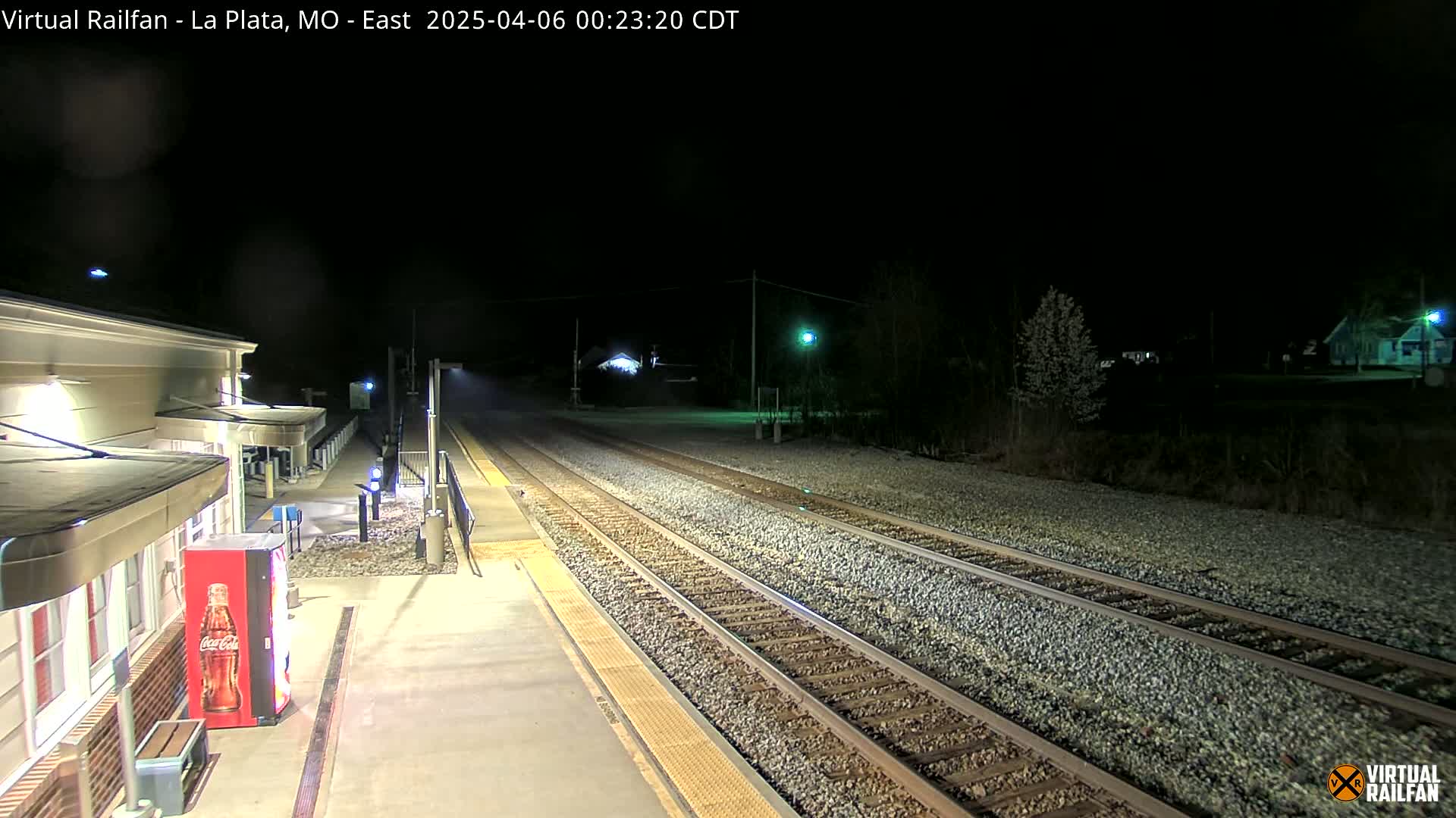 A nighttime view of a train station platform with a Coca-Cola vending machine, adjacent to train tracks extending into the darkness.